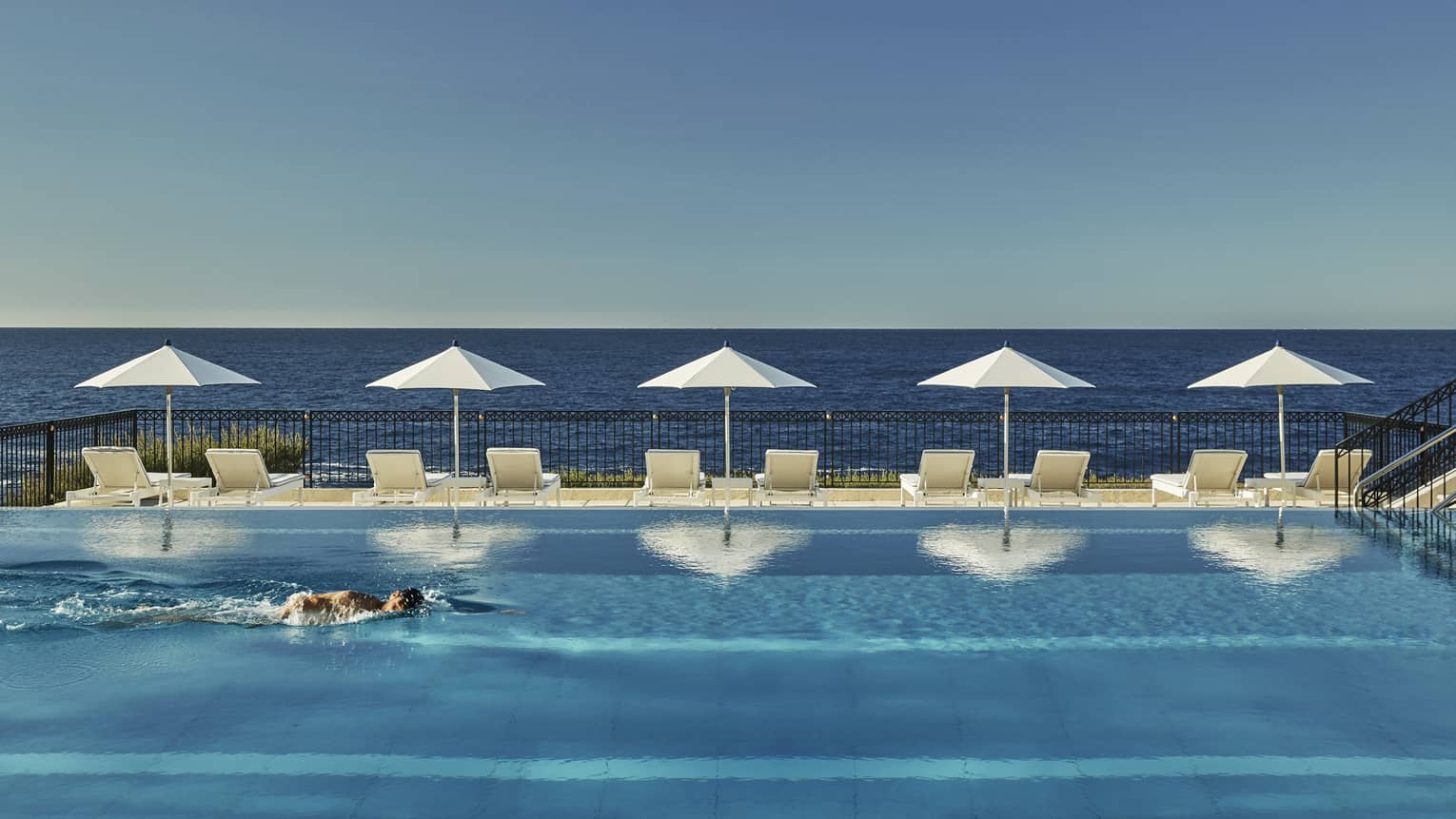 A man swims in outdoor lap pool, lined with lounge chairs and white umbrellas, looking out to sea