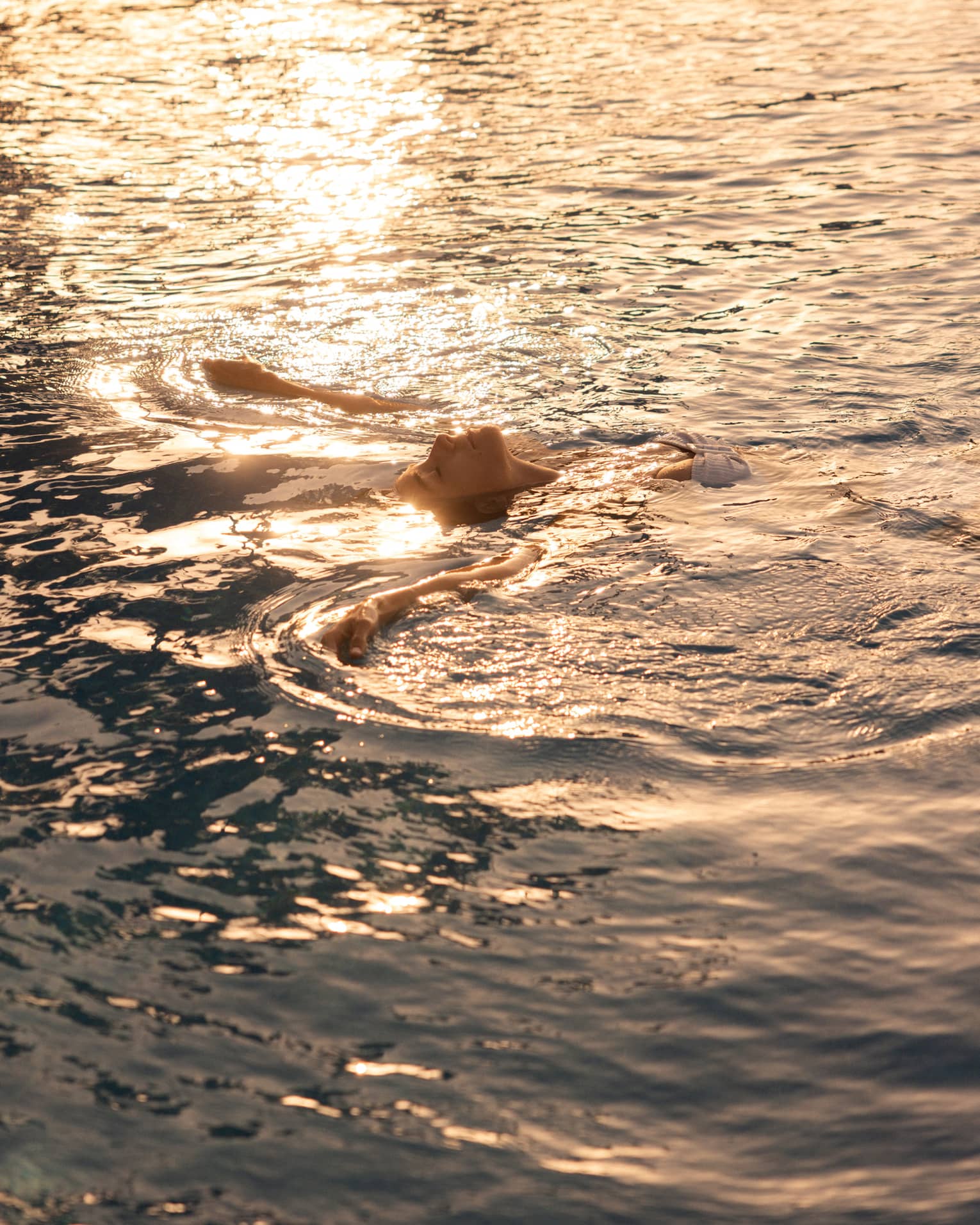 A person floating on their back in the ocean, with sunlight rippling across the water.