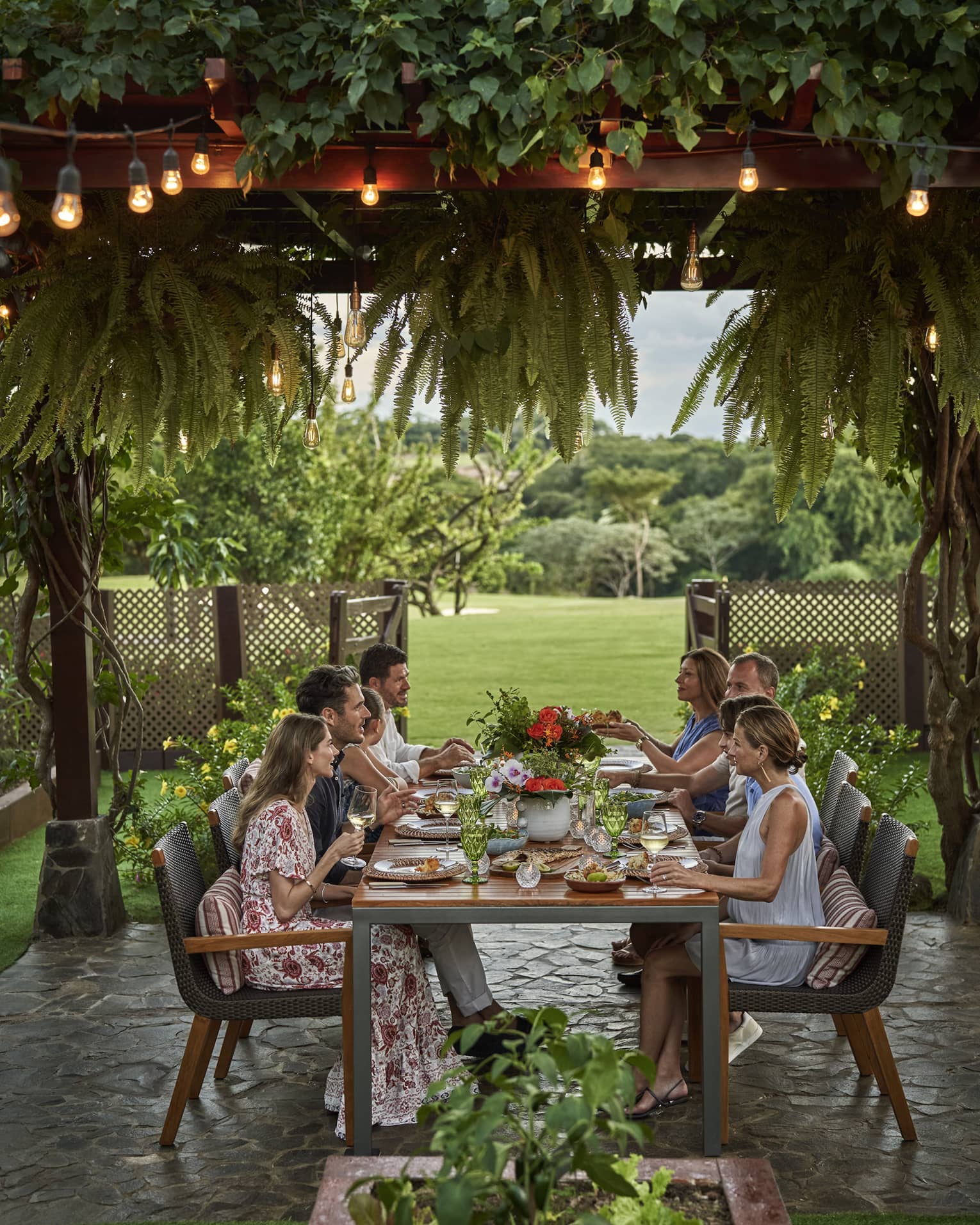 Eight people sit around a long dining table set up outside beneath a canopy of greenery and string lights