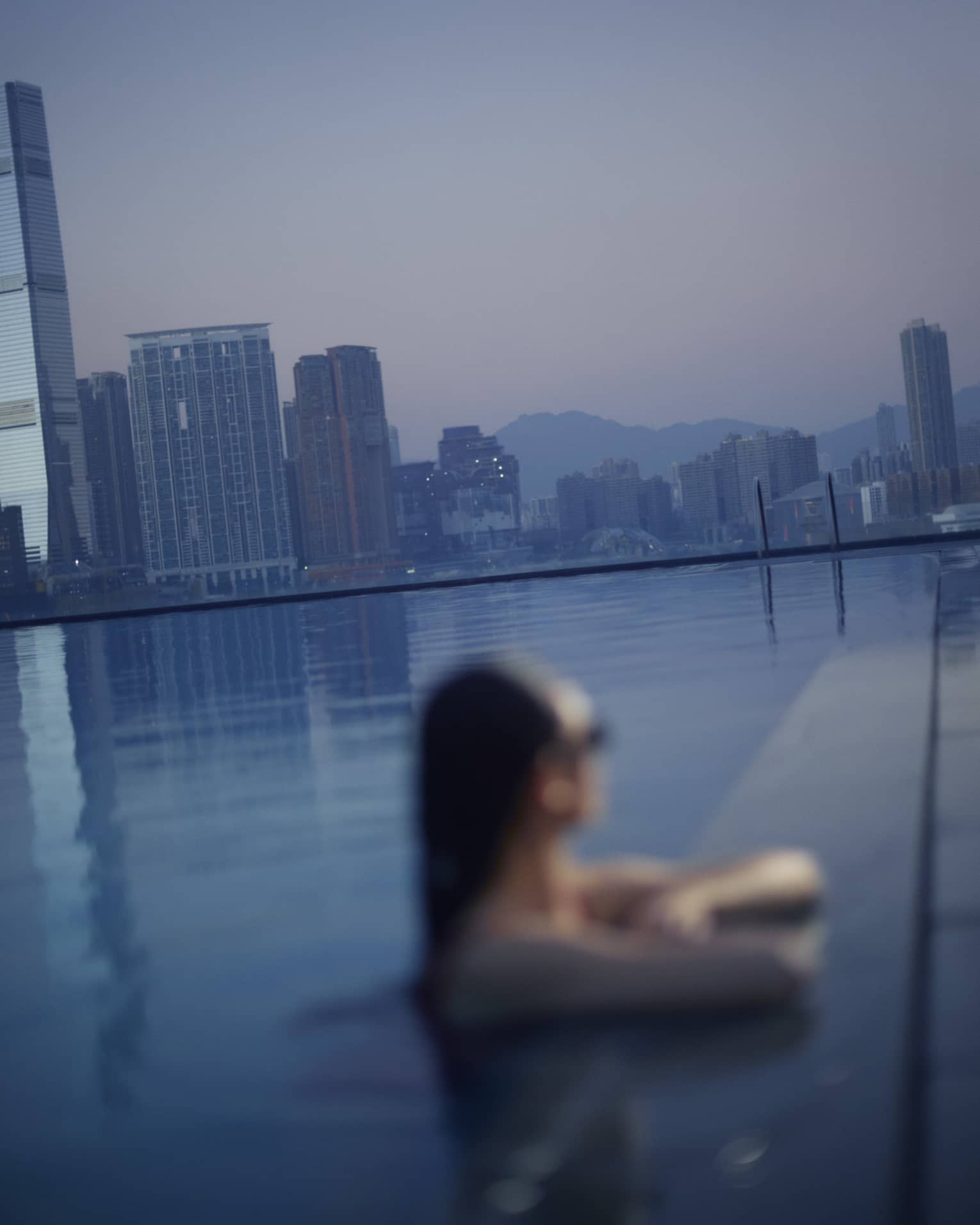 A woman in pool relaxes with elbows on pool deck, overlooking a city at dusk