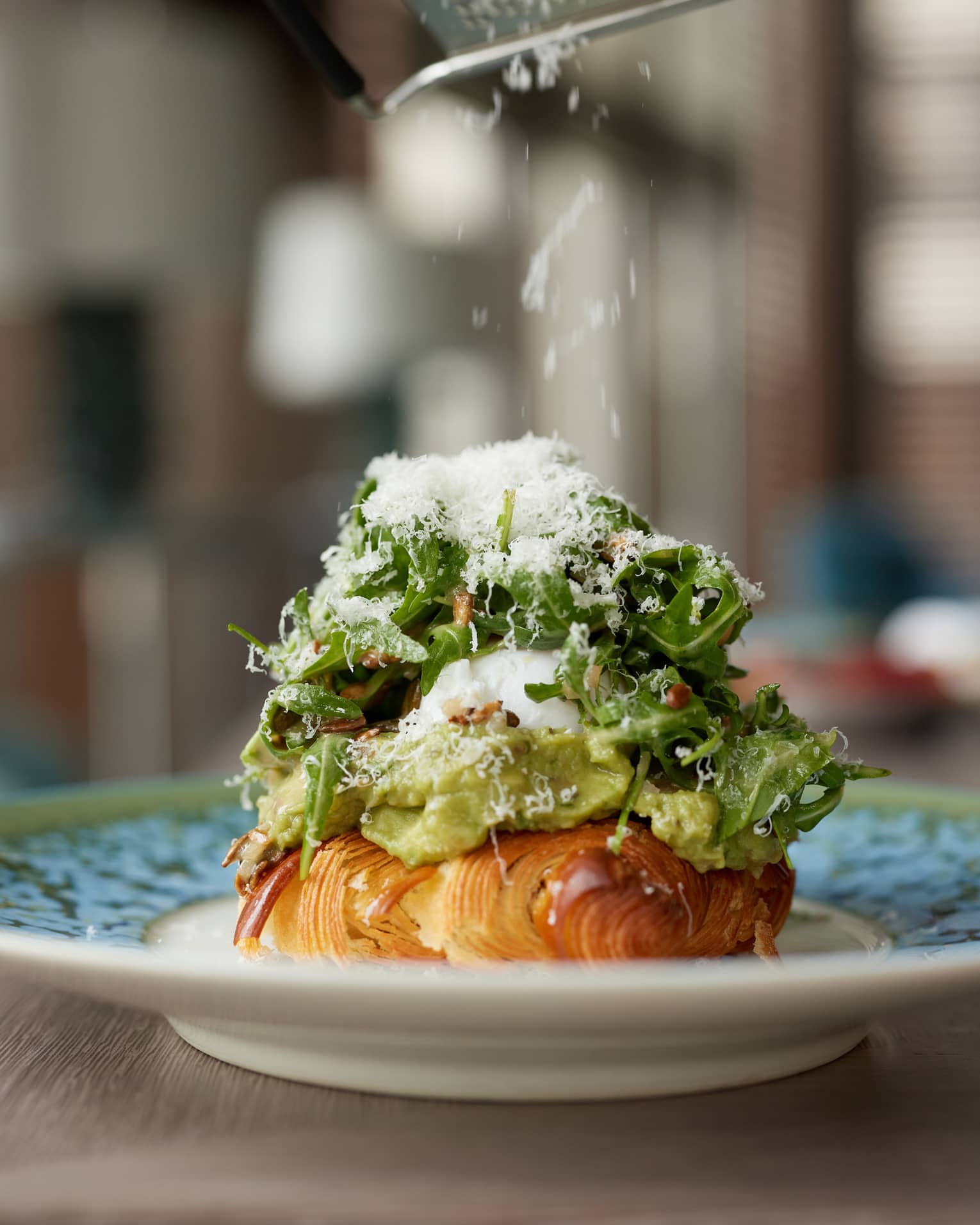 A close-up of a plate holding a croissant covered with avocado, egg and arugula salad topped with grated parmesan. 