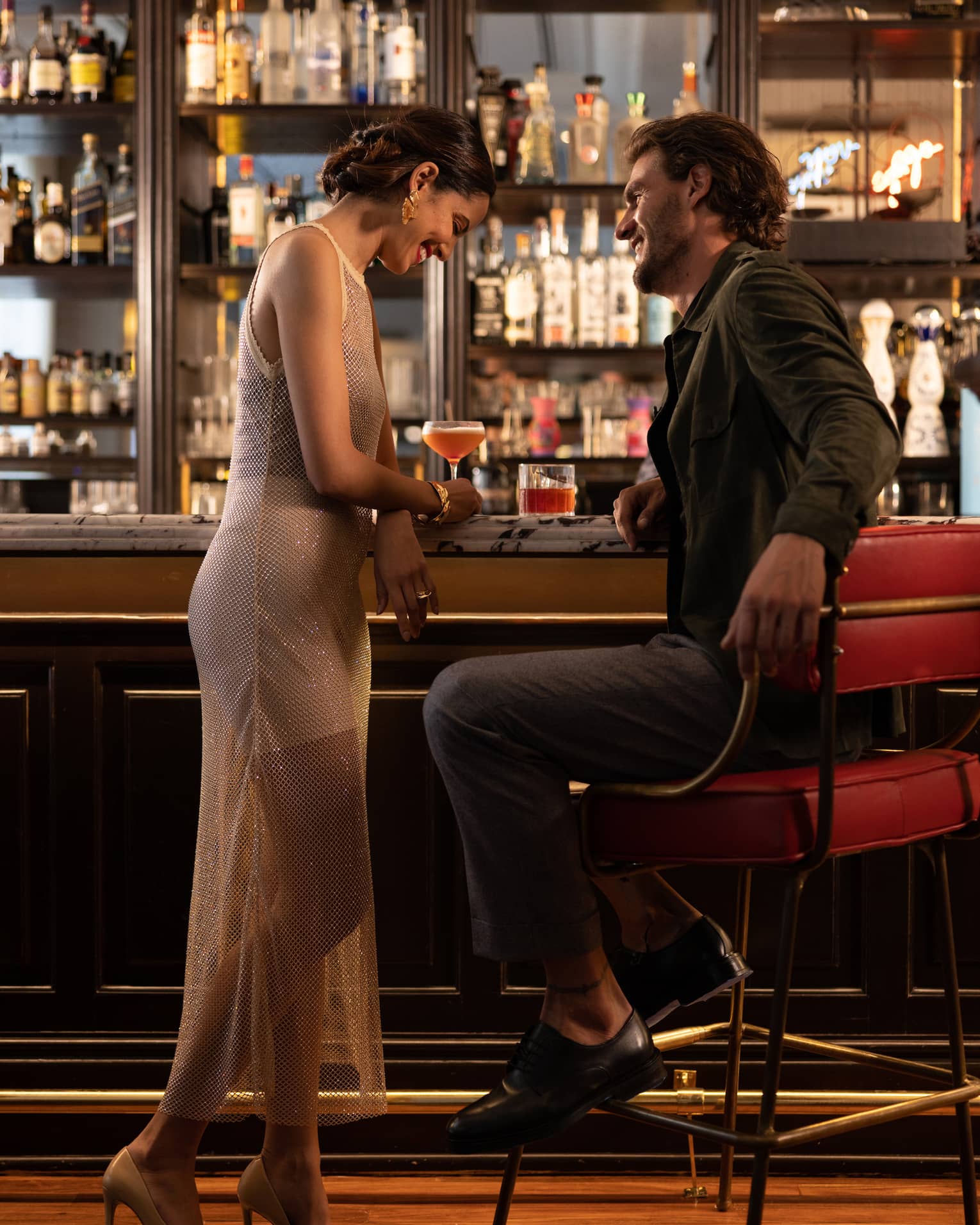 Elegantly dressed couple enjoy unique cocktails at a luxurious, dimly lit bar, with bottles of liquor on mirrored shelves. 