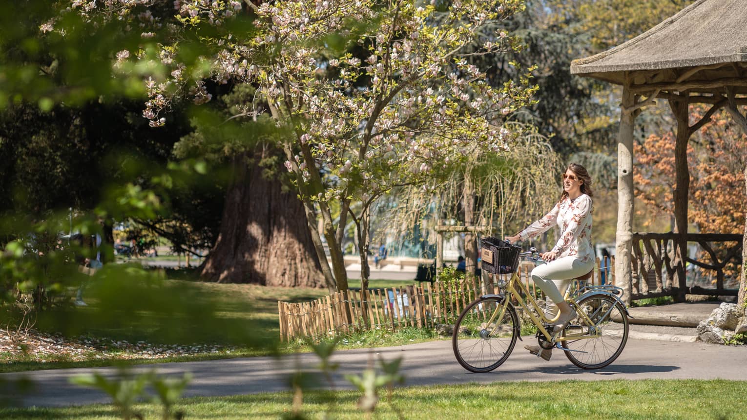Side view of a smiling guest on an bike with a basket coasting past mixed forest and spring blossoms in a sunny park.