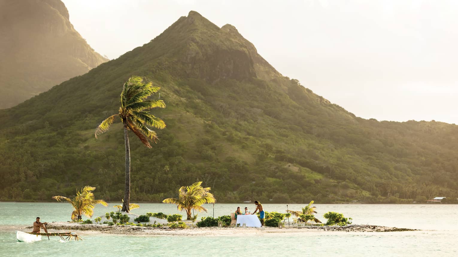 Couple dines on small private island with windswept palm trees in lagoon under mountain