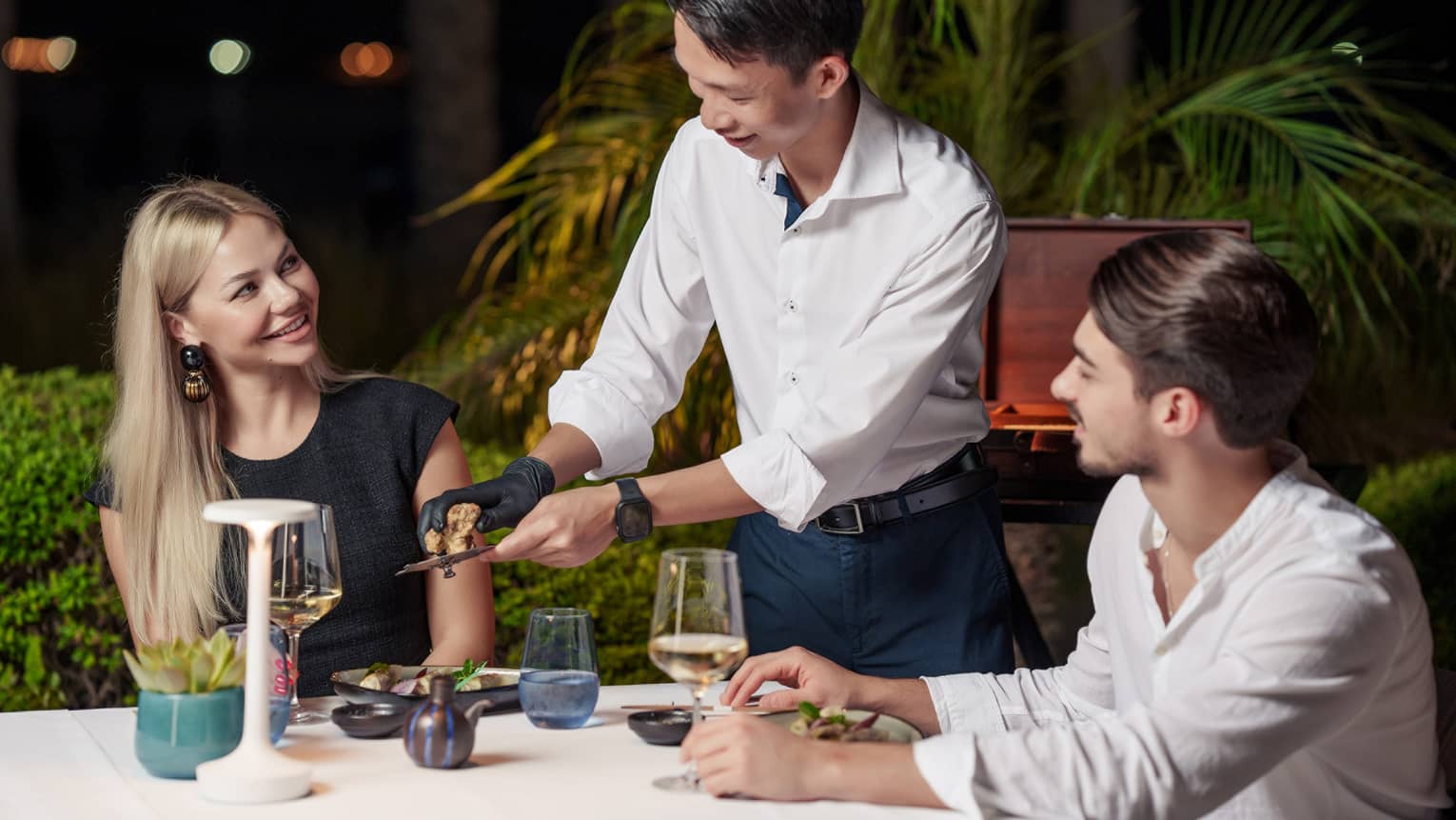 At a table for two amid foliage under the night’s sky, a server grates truffle over a diner’s dish as another diner looks on.
