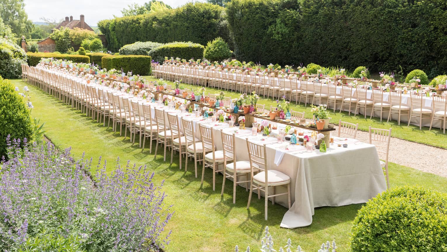 An outdoor reception in a garden, with very long rectangular tables set with flowers, plates and silverware