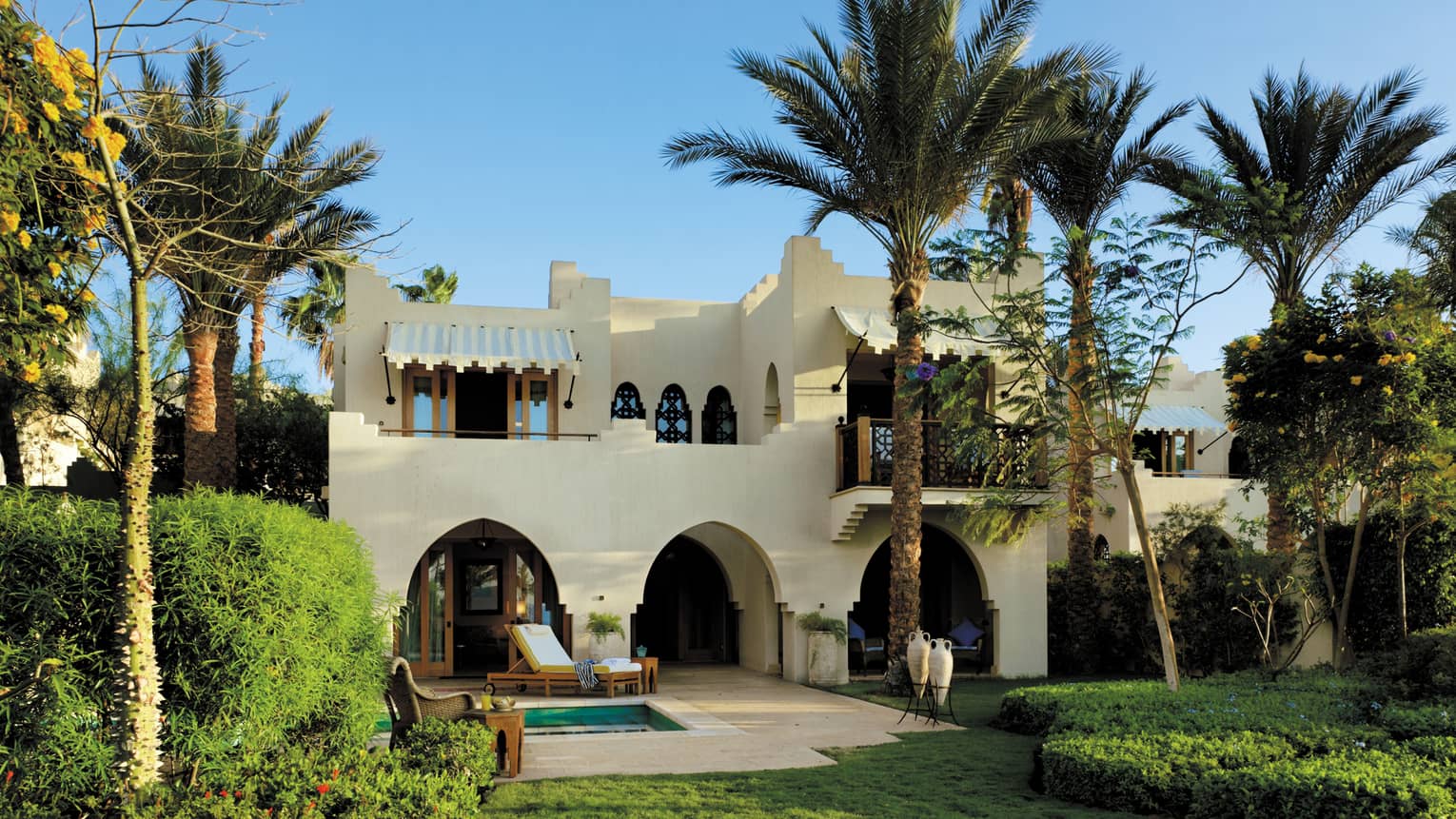 View across green lawn, garden to white two-storey luxury villa surrounded by palm trees