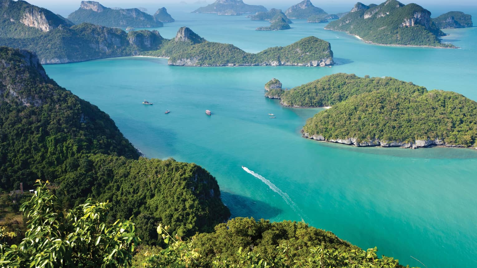 Aerial view of a dozen lush, forested, hilly islands of different sizes surrounded by calm turquoise water dotted with boats.