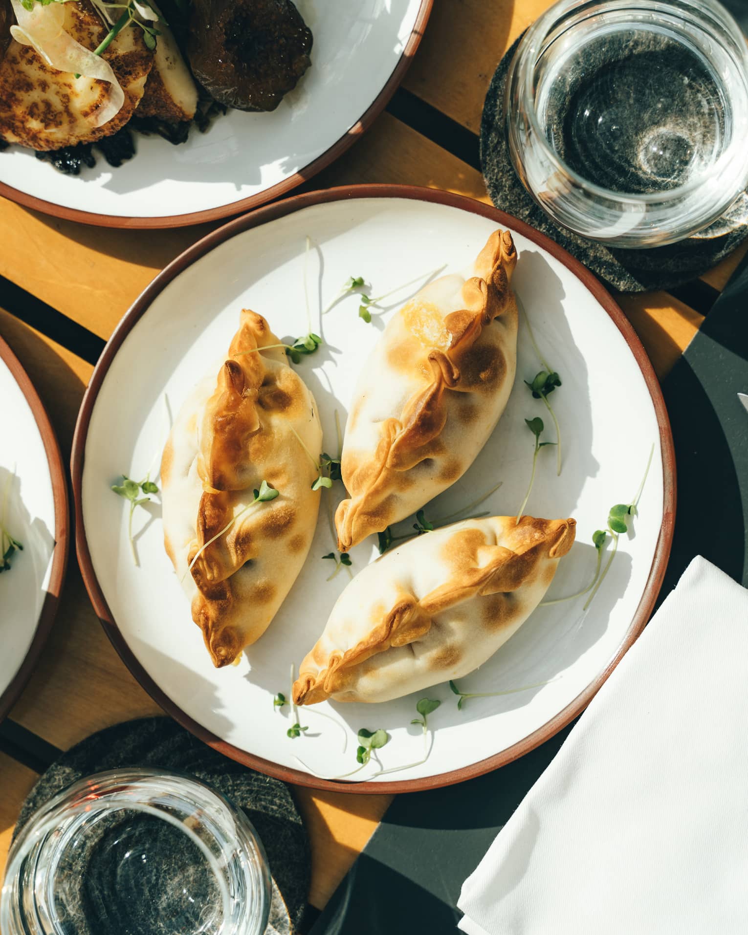 Three plump, golden-brown empanadas on a porcelain dish surrounded by sunlit water glasses and other plates of fine foods.