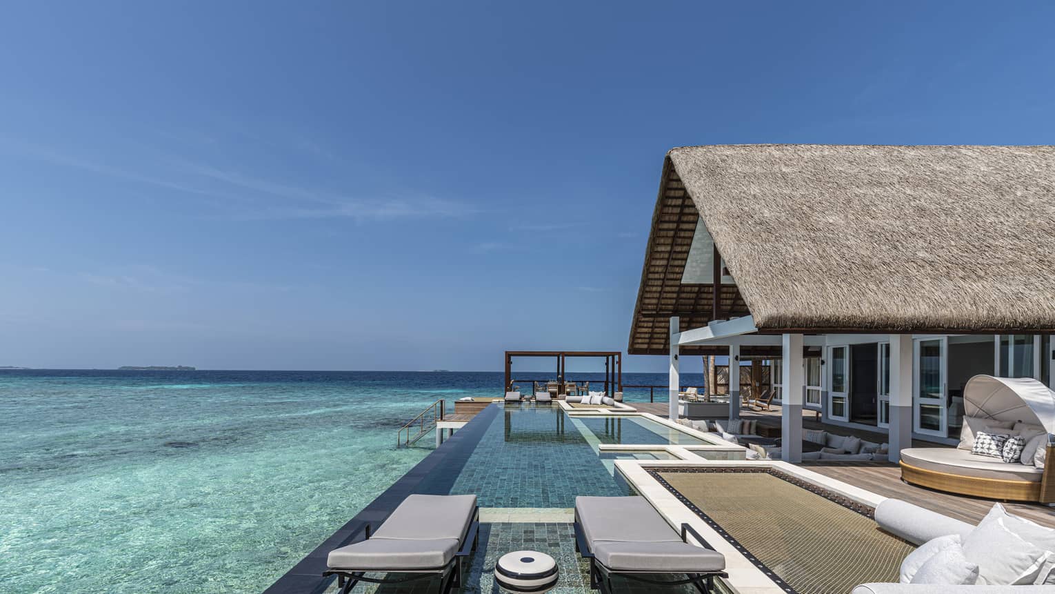 Modern overwater bungalow's expansive deck, with two chaise lounges and infinity pool