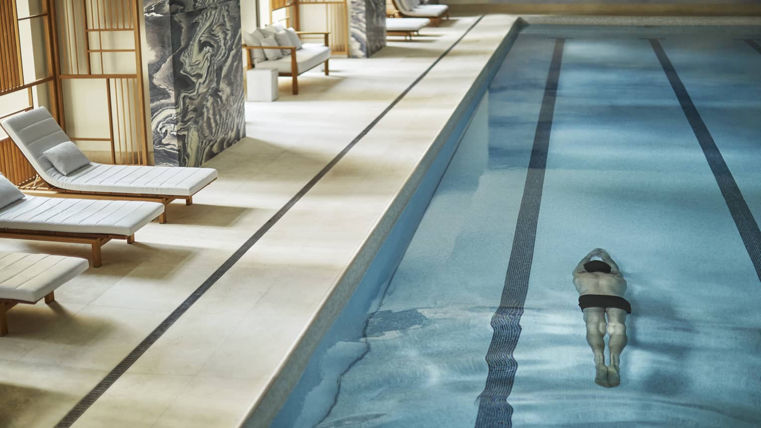 Man swims underwater in lane of indoor lap swimming pool by pool deck, chairs, marble wall