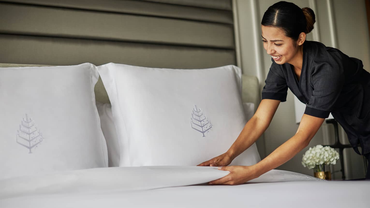 Hotel housekeeper smooths blanket under bed pillows with embroidered Four Seasons logos