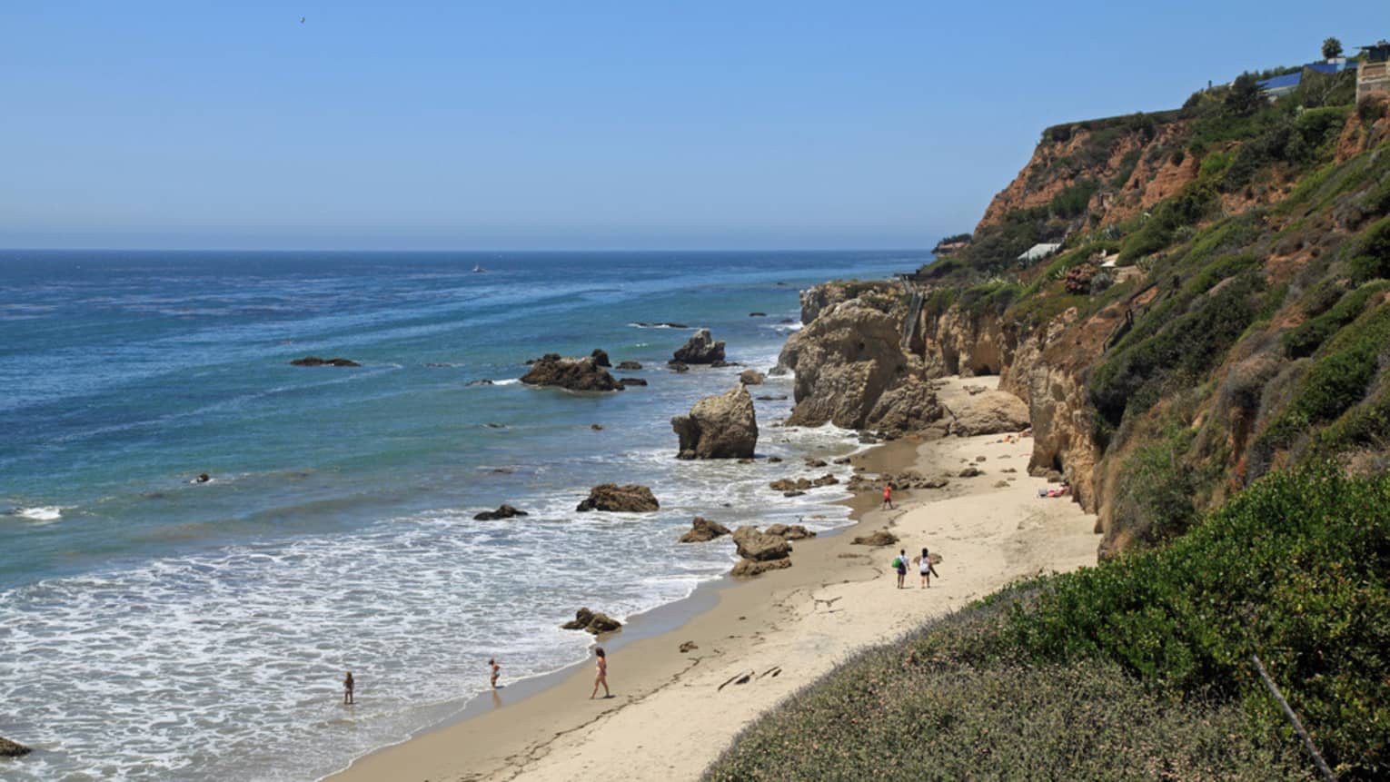 View of people on in ocean, walking on sand, rock formations and shrubbery 