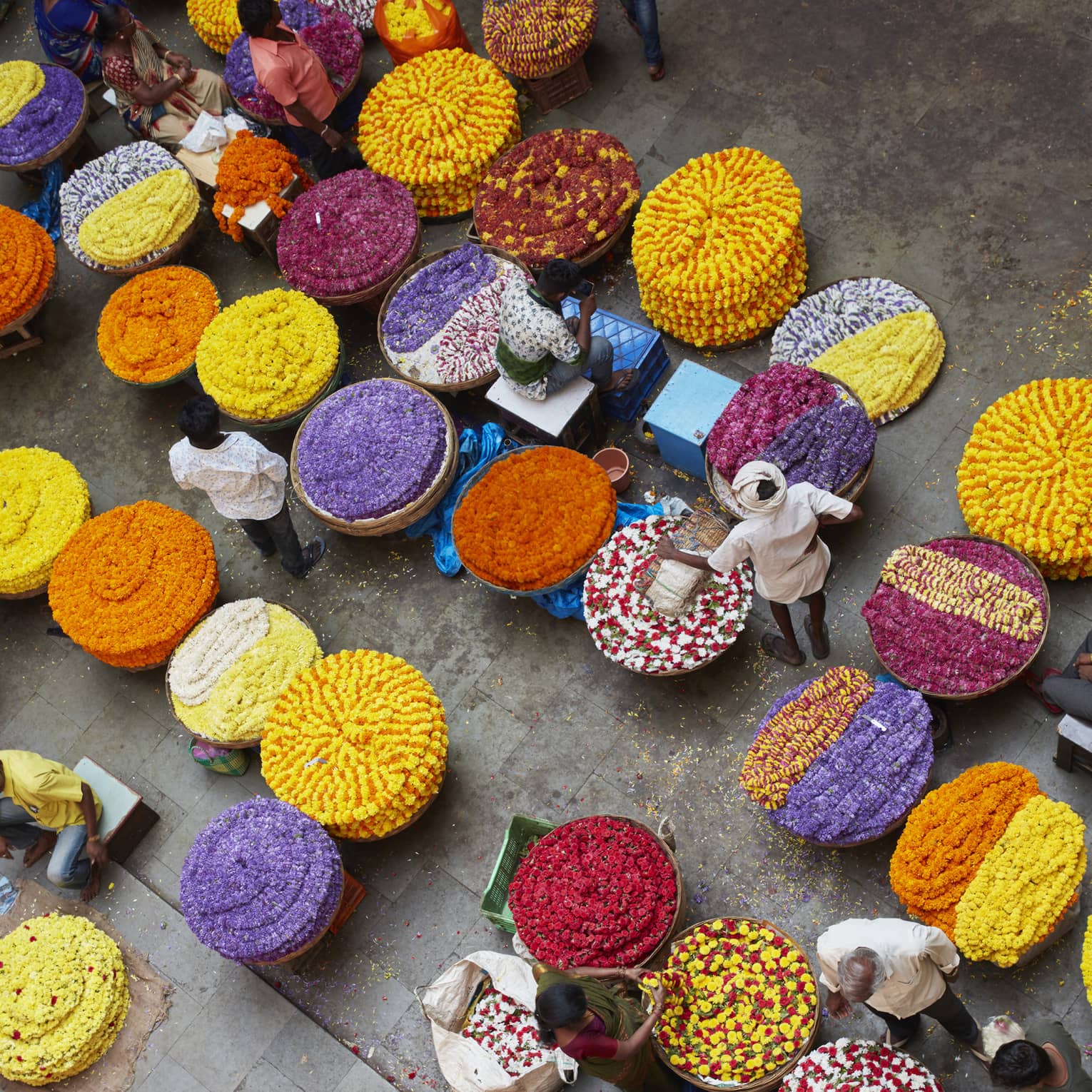 Bird’s-eye view of vendors with chains of vibrant, multicoloured flowers arranged in spirals in large round wicker baskets.