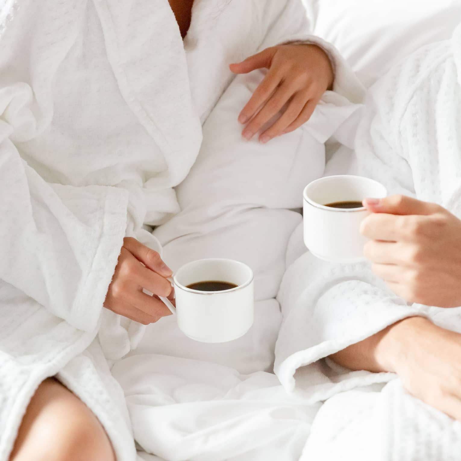 A faceless couple in white robes lounge on white bedding while holding mugs of black coffee.