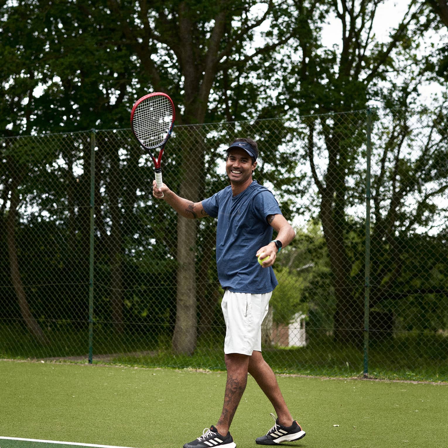 Person standing and smiling while readying to serve on green tennis court with fence and trees in backdrop