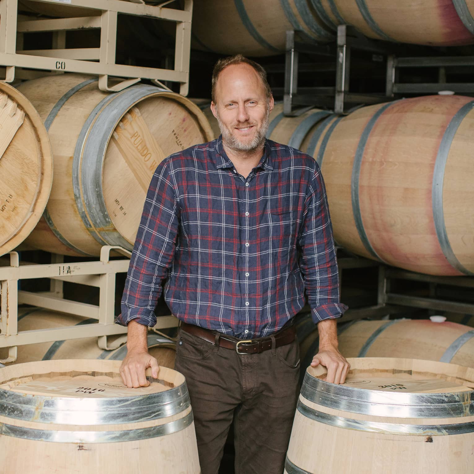 Thomas Brown stands before two racks of wine barrels as he rests his hands on two upright barrels and smiles at the camera.