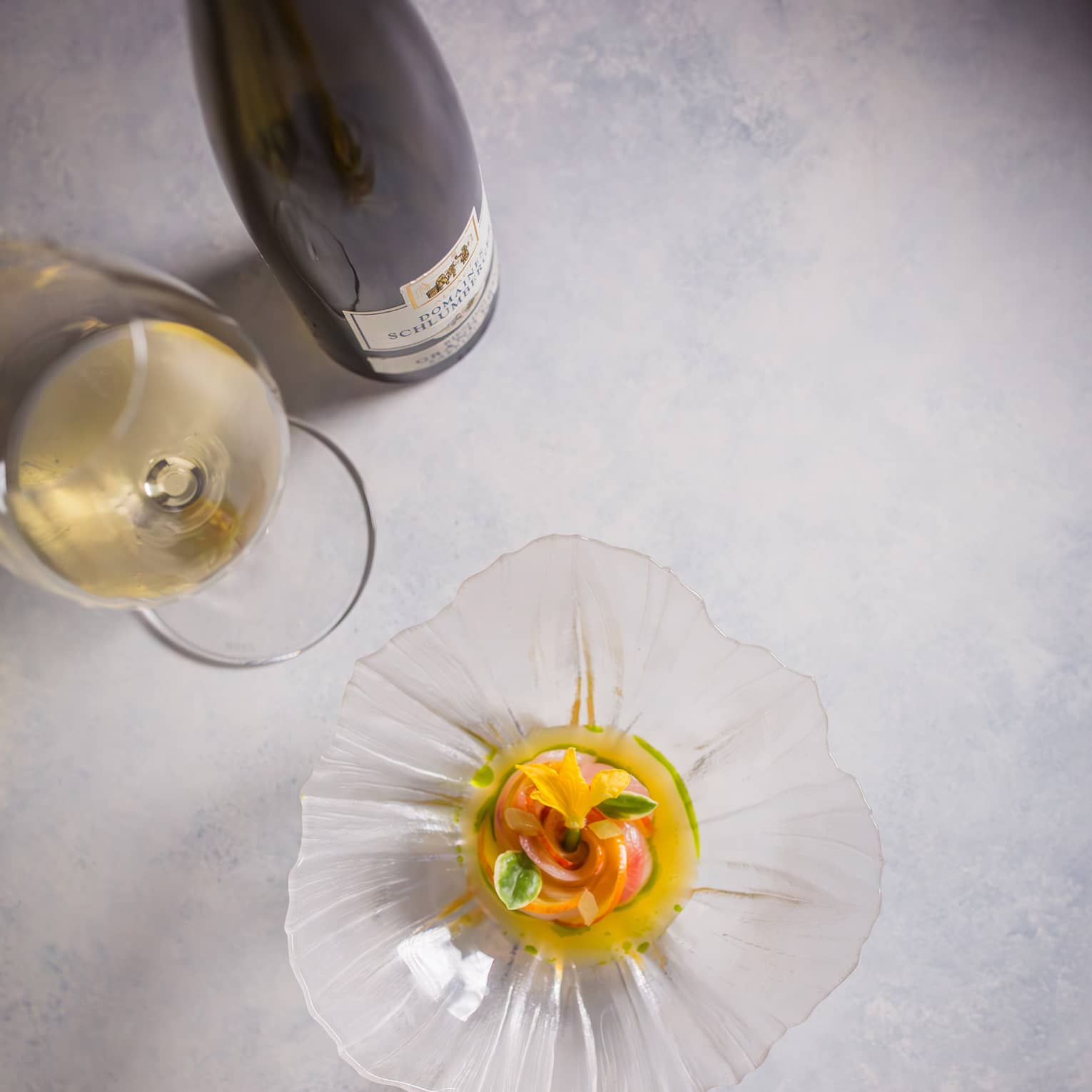 Overhead view of a colourful appetizer garnished with florals in a glass, flower-shaped dish beside a glass of white wine.