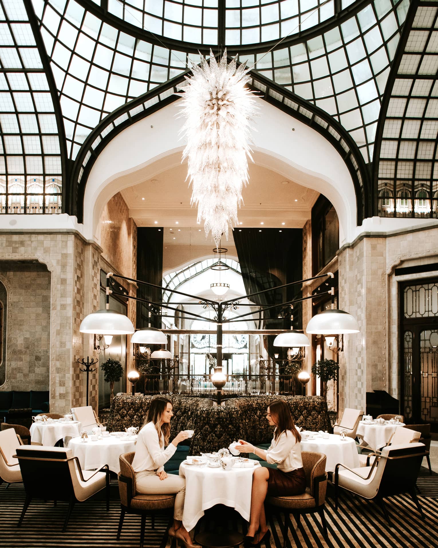 Elegant dining area with a grand chandelier, high arched glass ceiling, and modern seating, where two people are enjoying tea at a small table.
