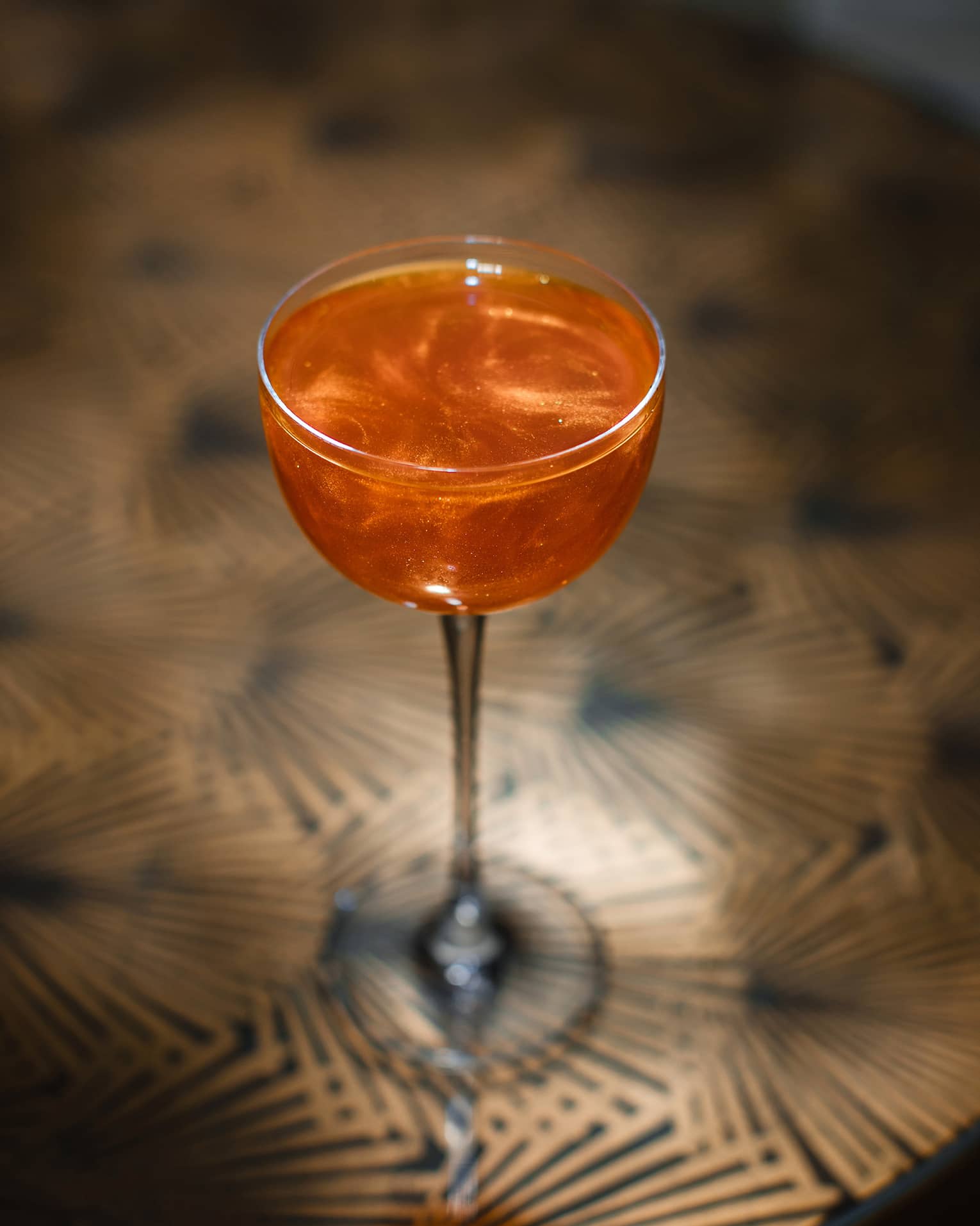 Bright-orange clear cocktail in wine glass atop bronze patterned table