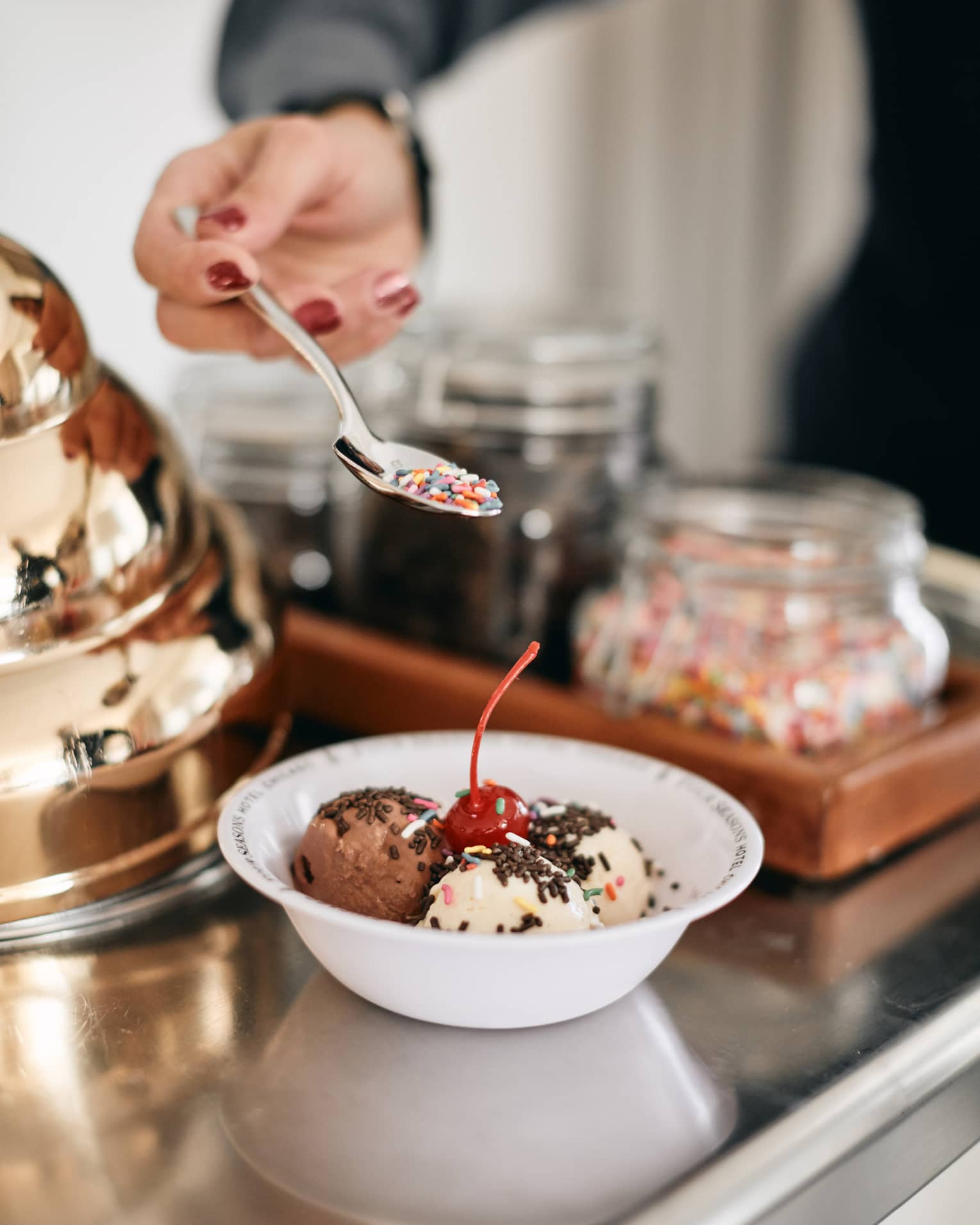 A member of staff creating a colorful ice cream bowl for a guest, there are various flavours in the bowl, sprinkles, and a cherry.