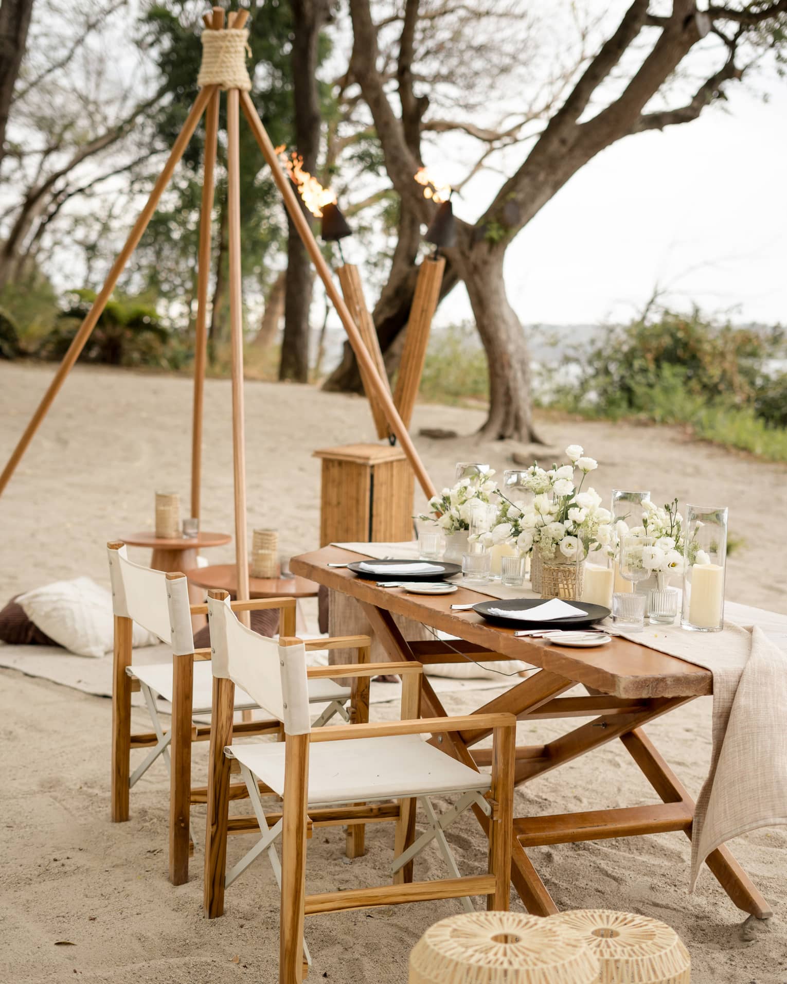Wooden dining table and chairs set up on a beach, decorated with white floral centrepieces and candles