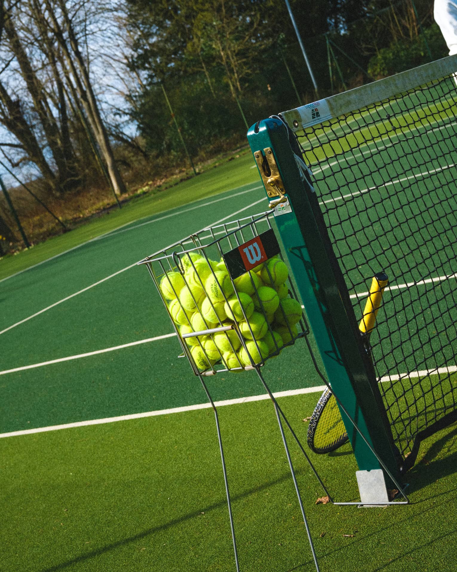 Tilted close-up of a basket filled with yellow tennis balls beside a net upon which leans a racquet on a green court.