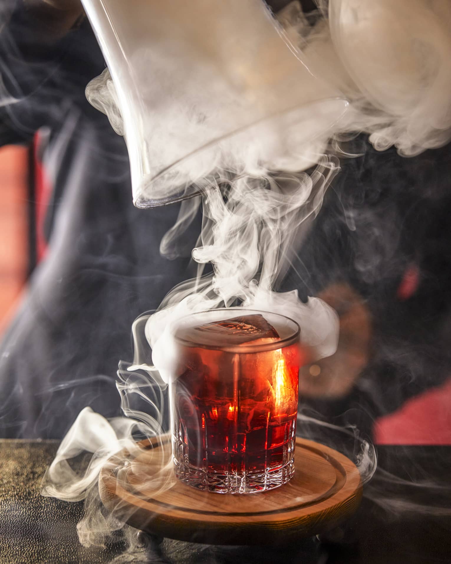 Bartender swirls smoke on top of a shot glass of amber-coloured liquor 