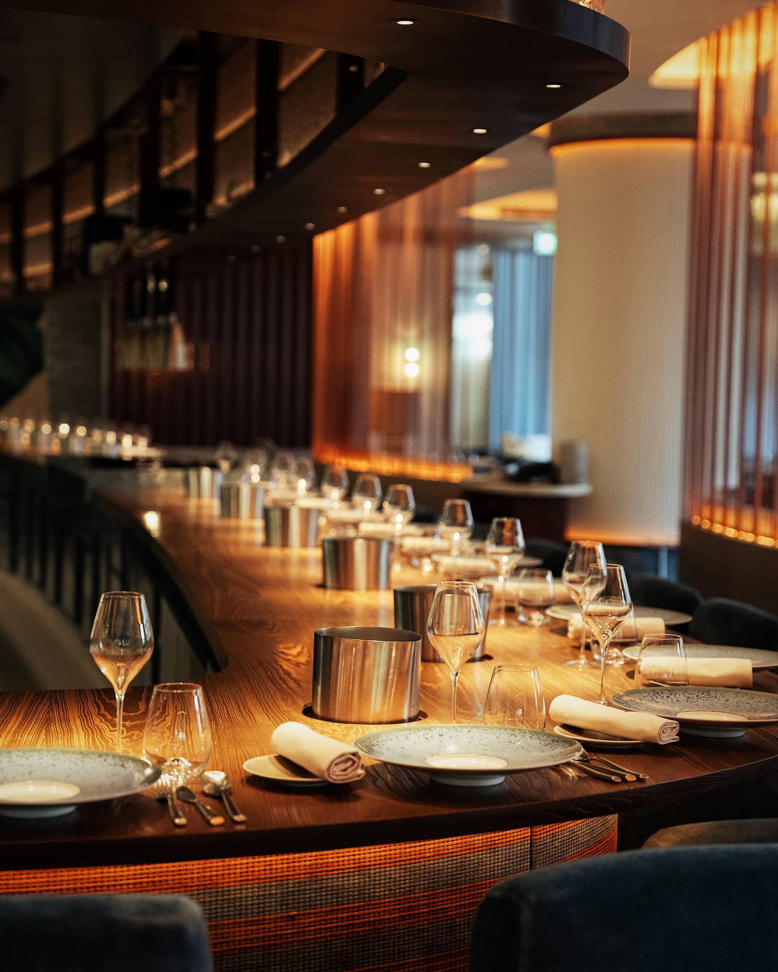 A long, curved solid wood bar with a dozen place settings in warm, golden lighting.