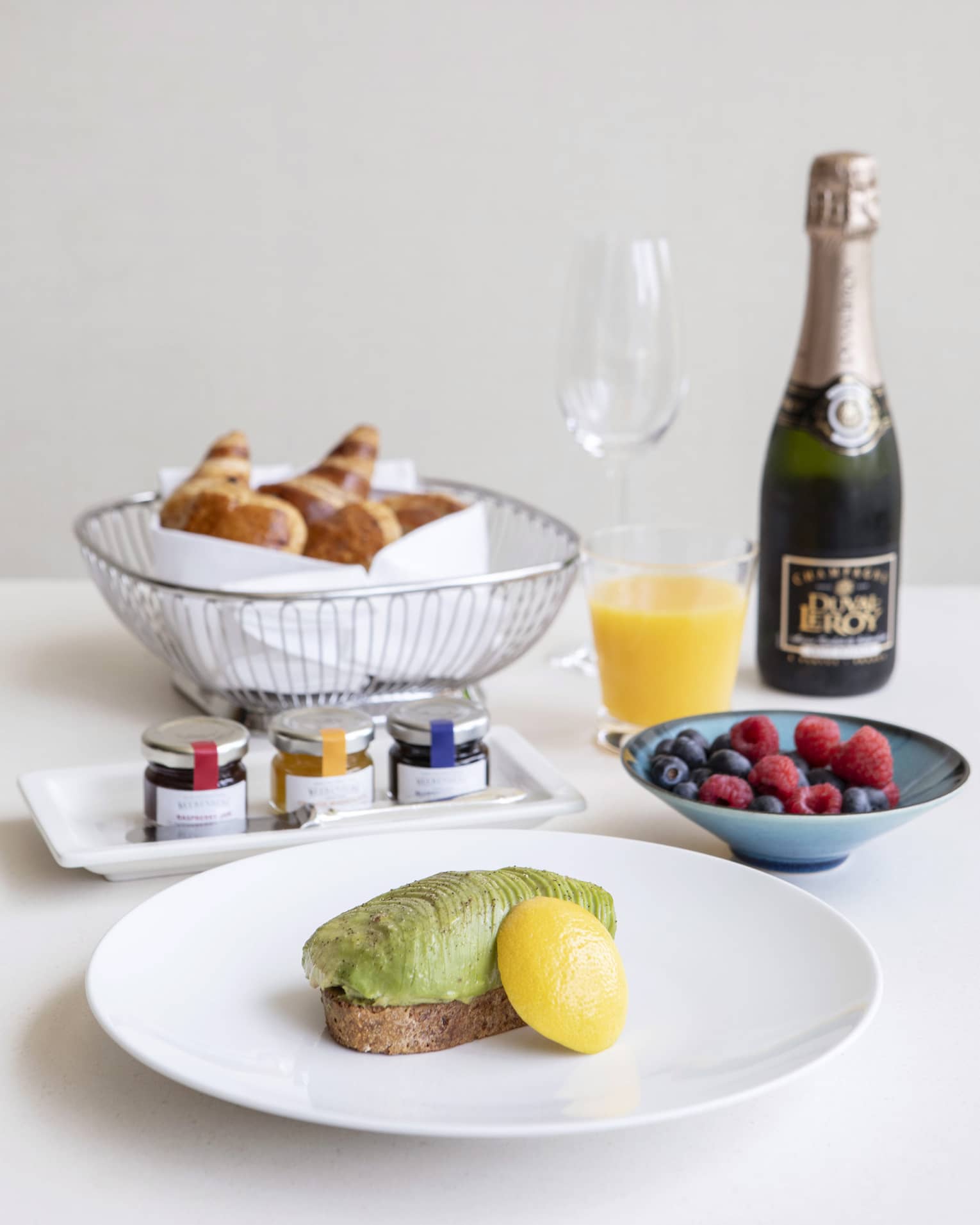 Luxurious breakfast including avocado toast, a basket of croissants, fresh fruit, and champagne and orange juice.