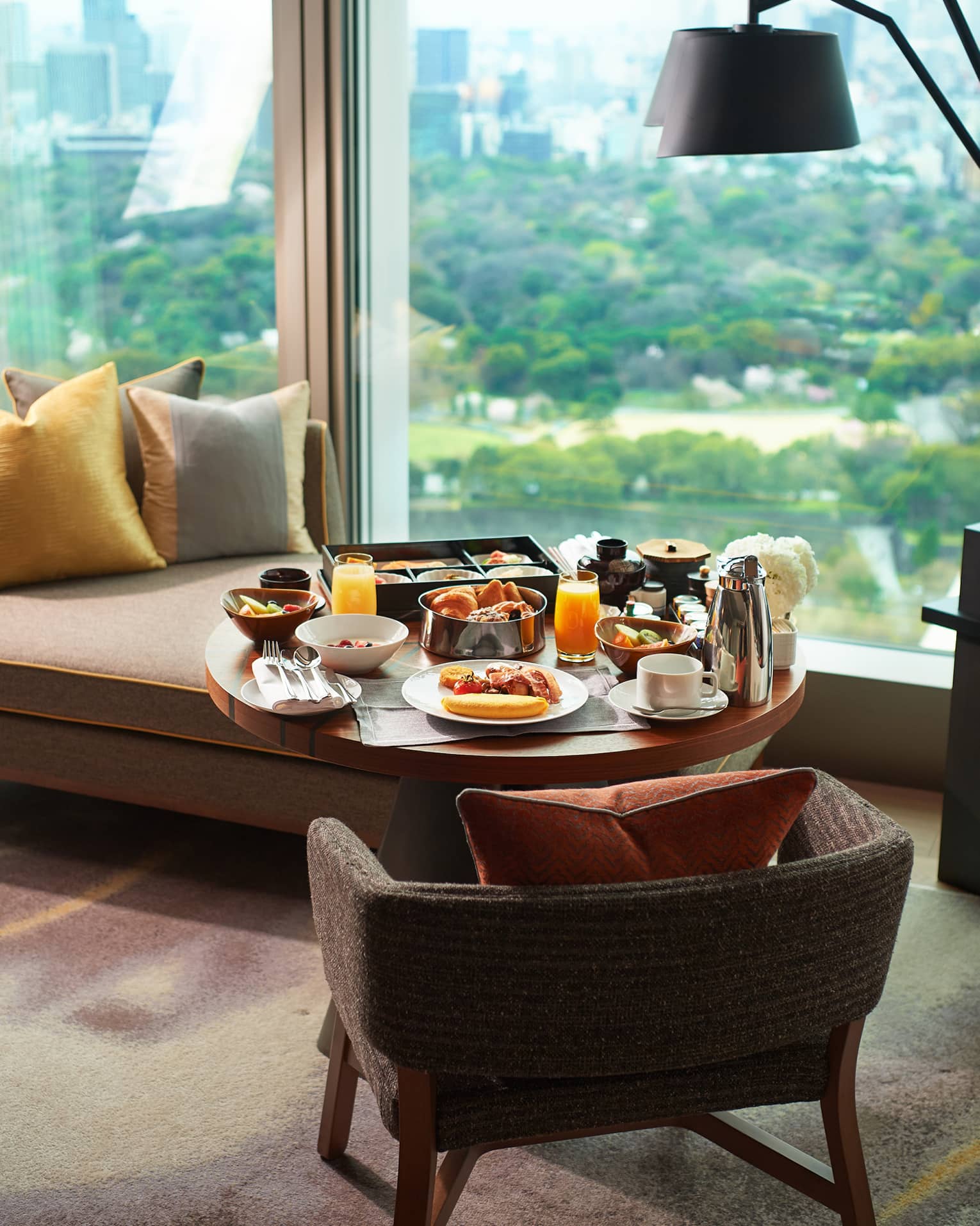A breakfast tray of pastries, fresh fruit, juice and coffee on a guest room lounge chair at a window overlooking lush gardens.