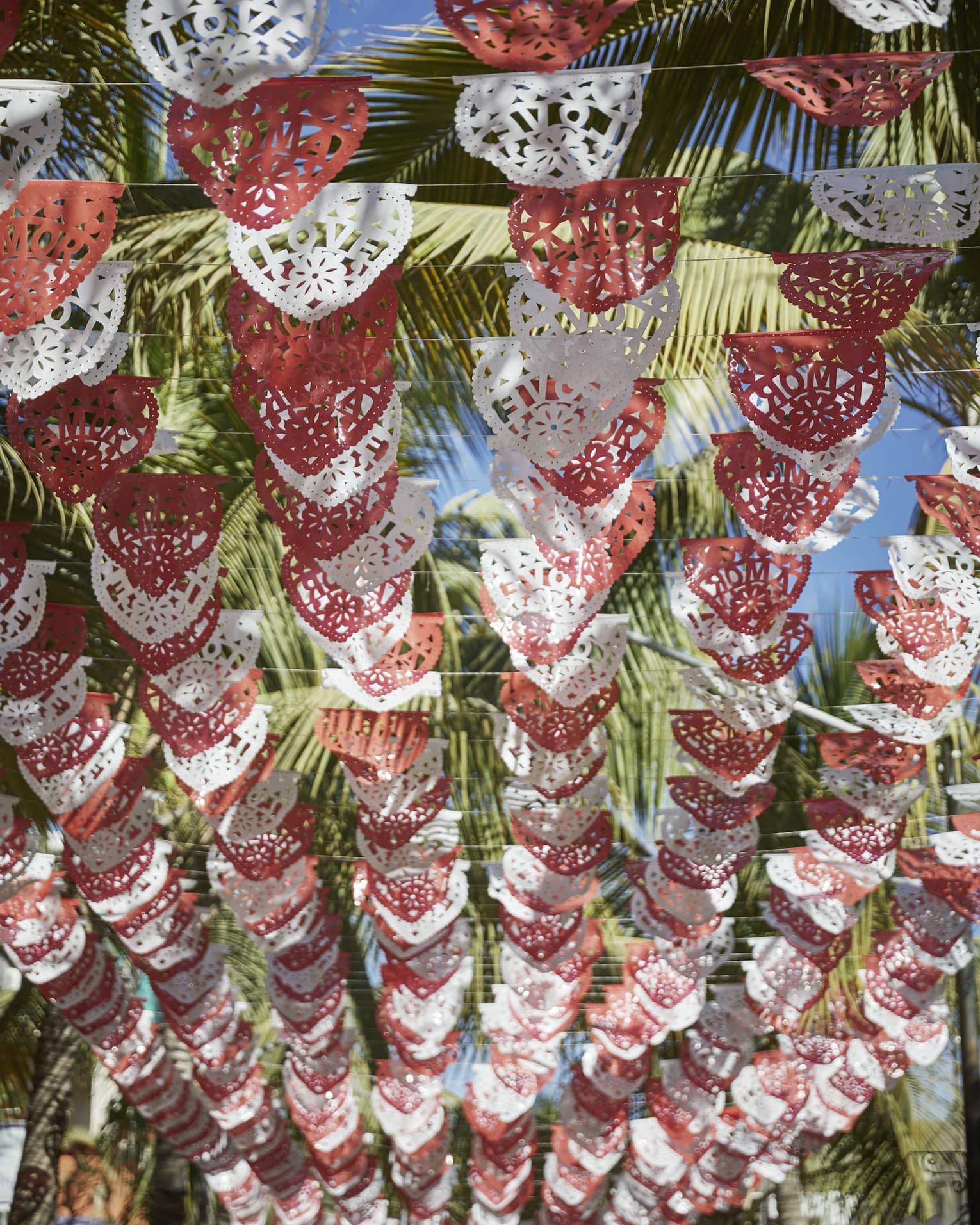 Looking up at rows of red and white Mexican Papel Picado banners hanging above, with palm trees and a sunny sky above them.