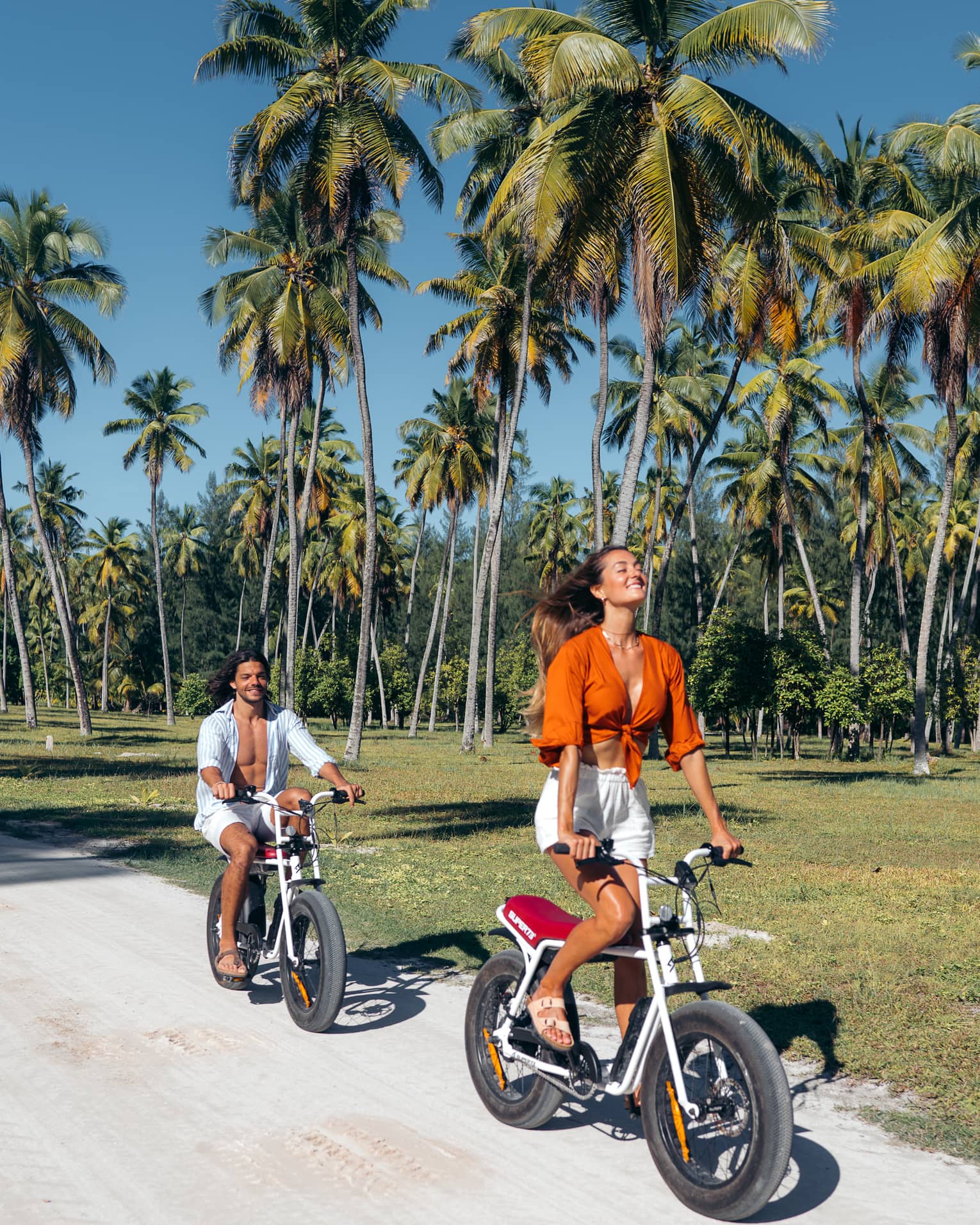 Smiling, eyes closed, face tilted toward the sky, a guest rides an e-bike, a friend trailing, rolling past palm trees. 