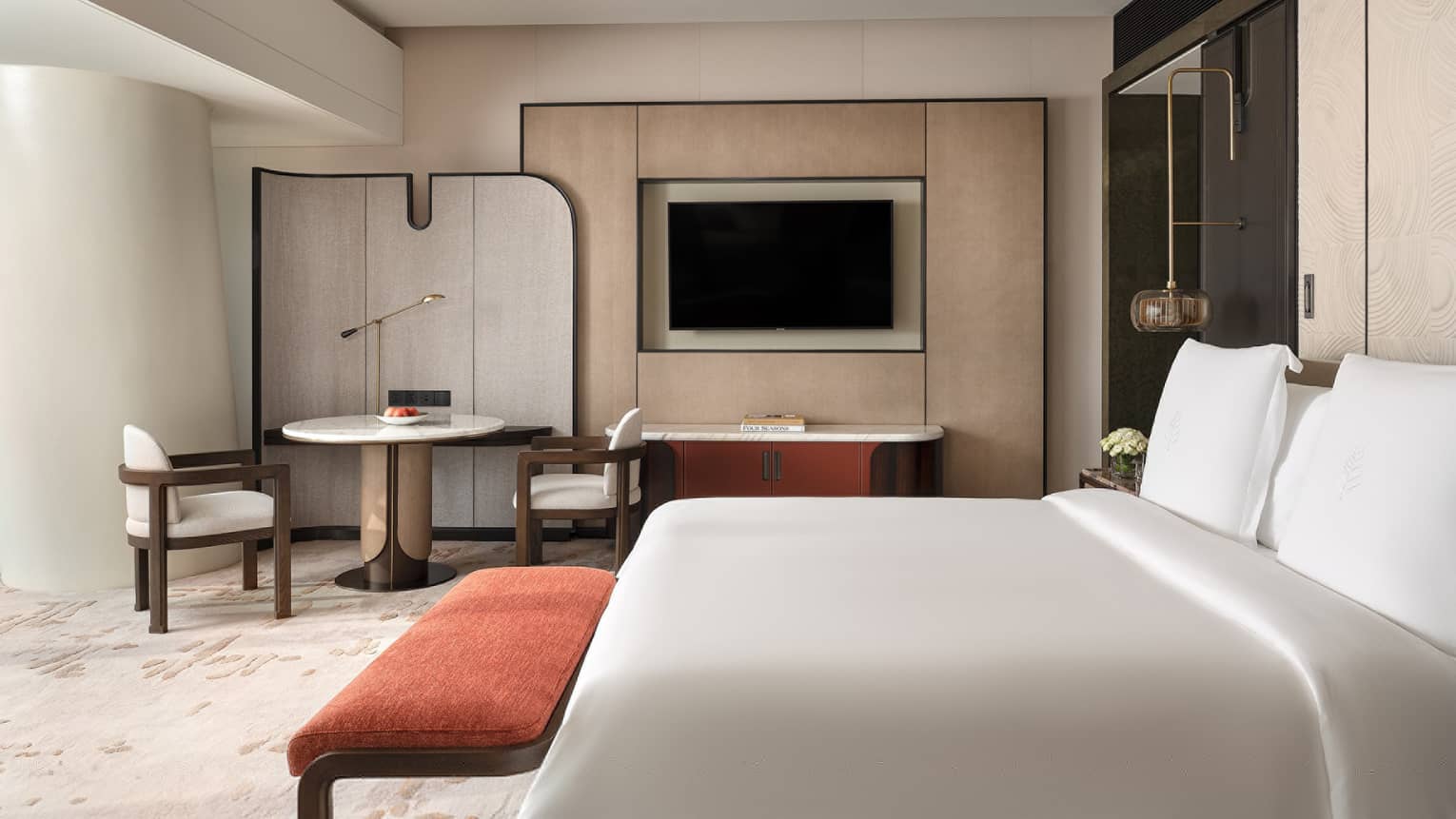 Contemporary hotel room with king bed, red cushioned bench, round table with two chairs and TV
