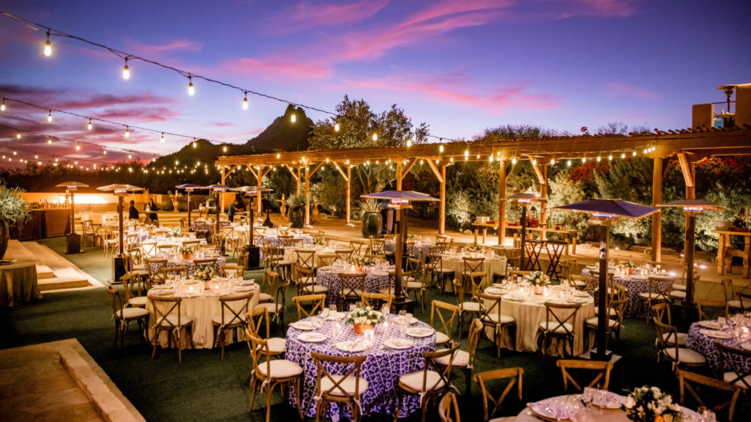 A large outdoor area with many round tables and wood chairs surrounded by small lights hanging on wires.