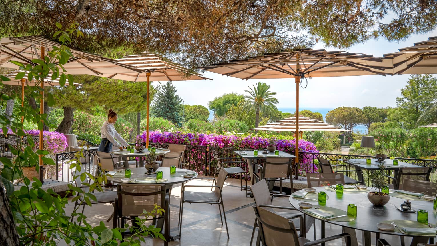 Waiter sets a table on outdoor restaurant terrace, surrounded by greenery and magenta-coloured flowers