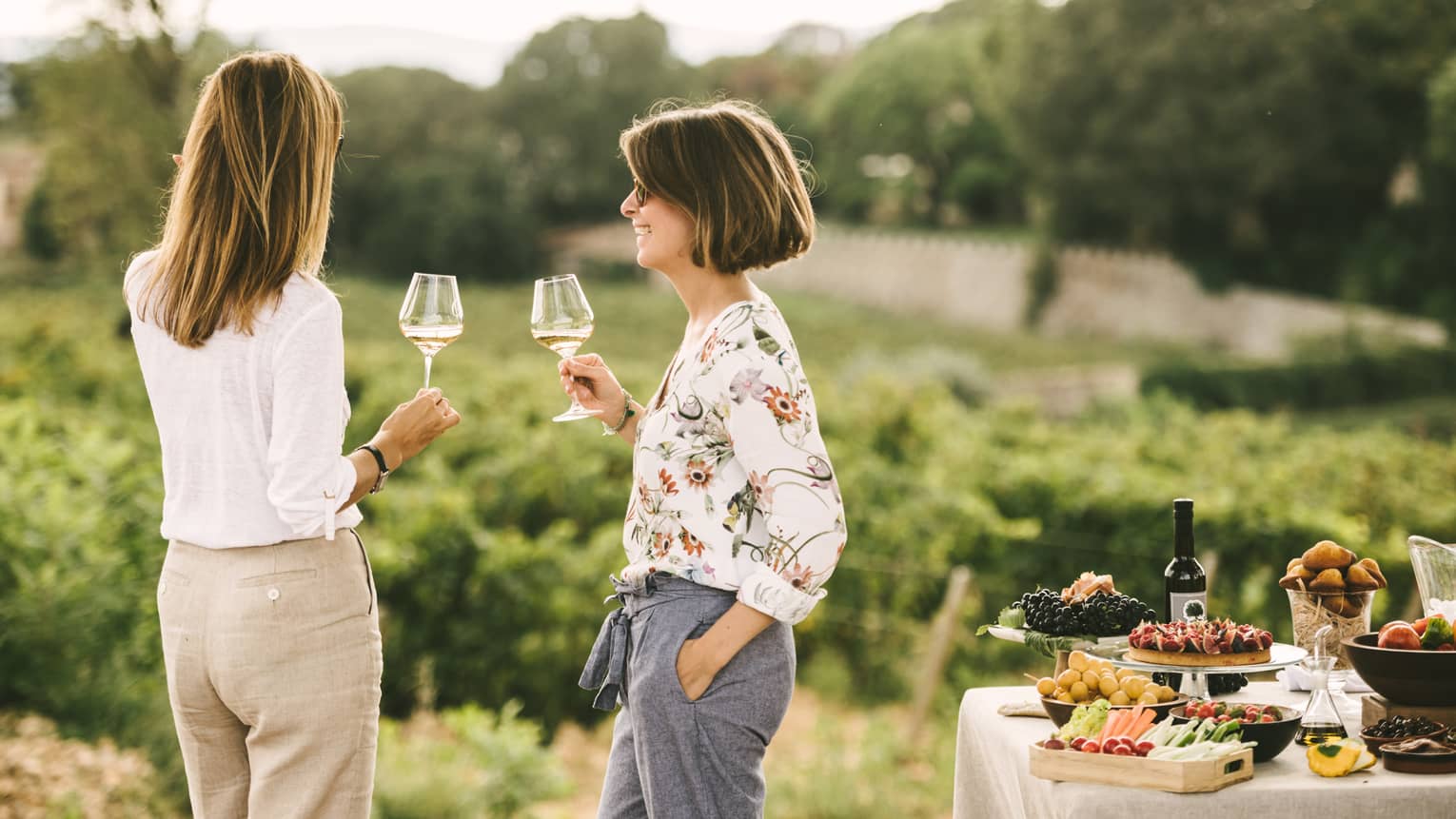 Two women drink a glass of white wine while standing among vineyards and private picnic