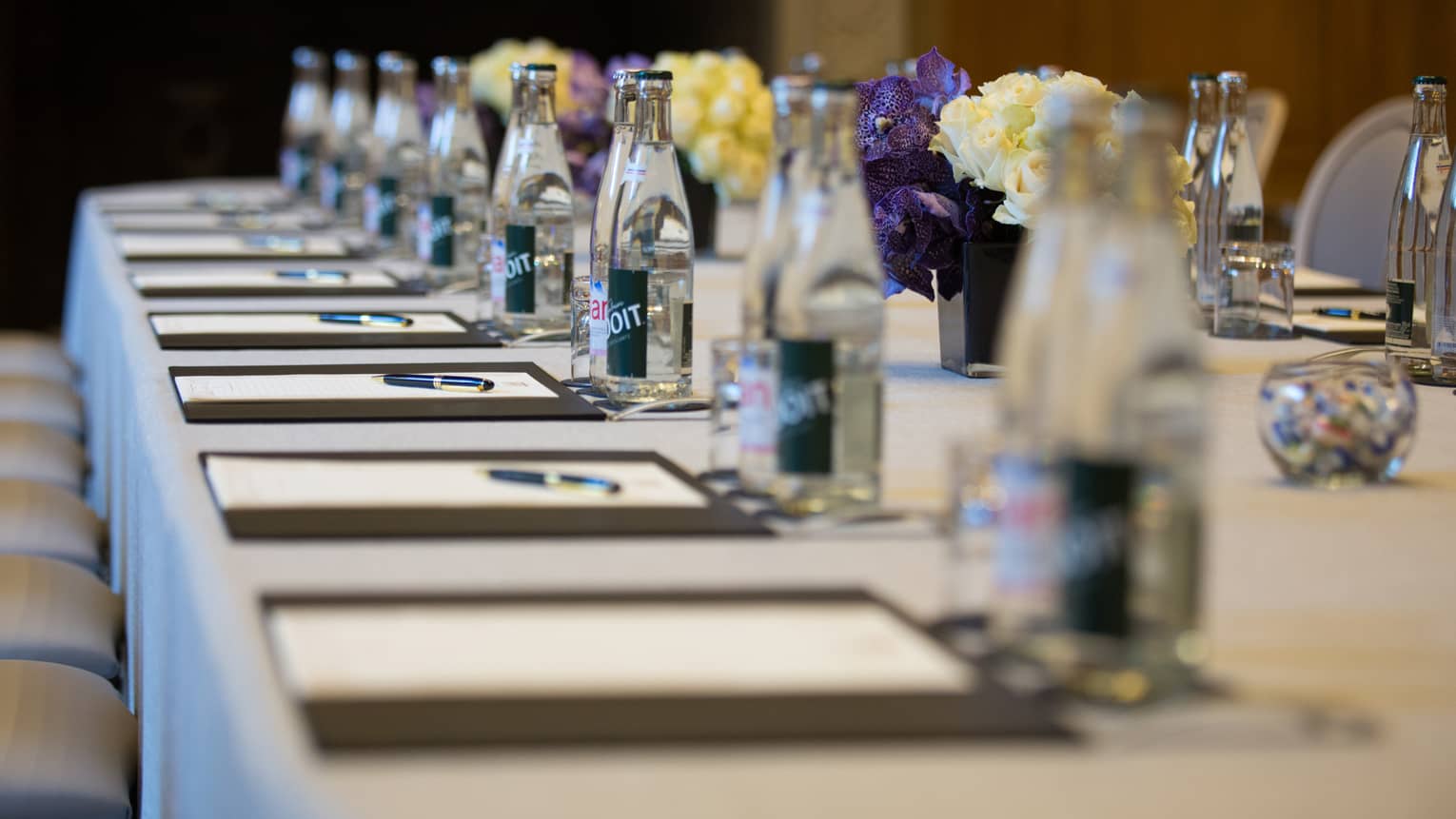 Row of meeting agendas, glass bottles, small vases with flowers on boardroom table