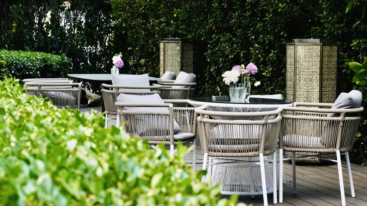Flanked by tall hedges, two tables surrounded by beige, cushioned rattan chairs hold glass vases of pink and white flowers.