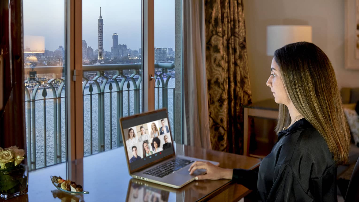 Woman working on a computer in a guest room, with city skyline view through the window.