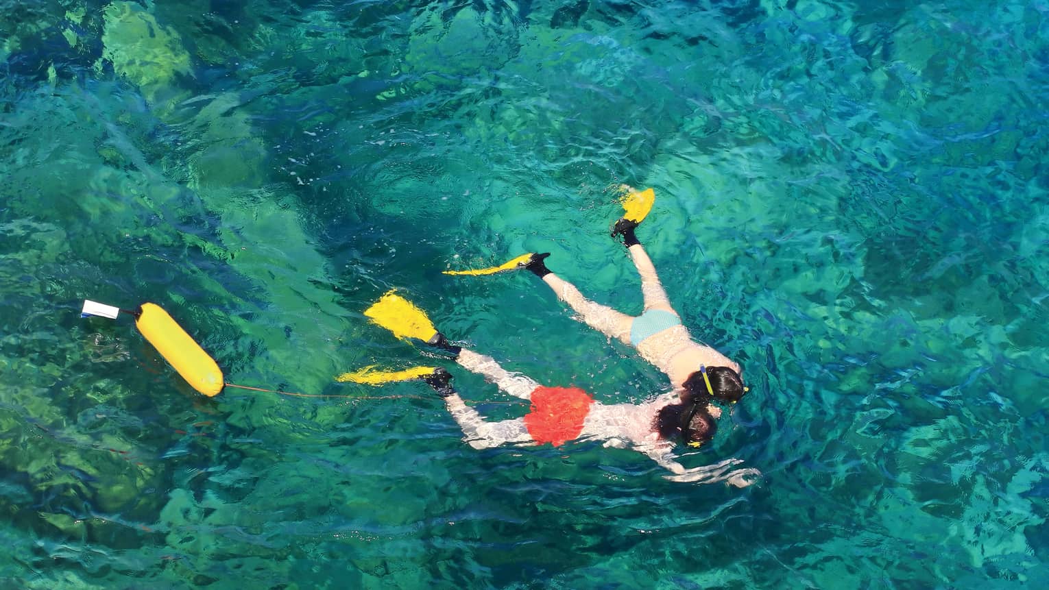 View from above of two guests in flippers snorkelling hand in hand, surrounded by crystal turquoise waters.