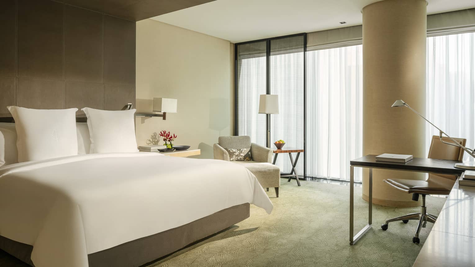 Guest room featuring a king bed, armchair, workspace, side table with red flowers and floor-to-ceiling windows with curtains