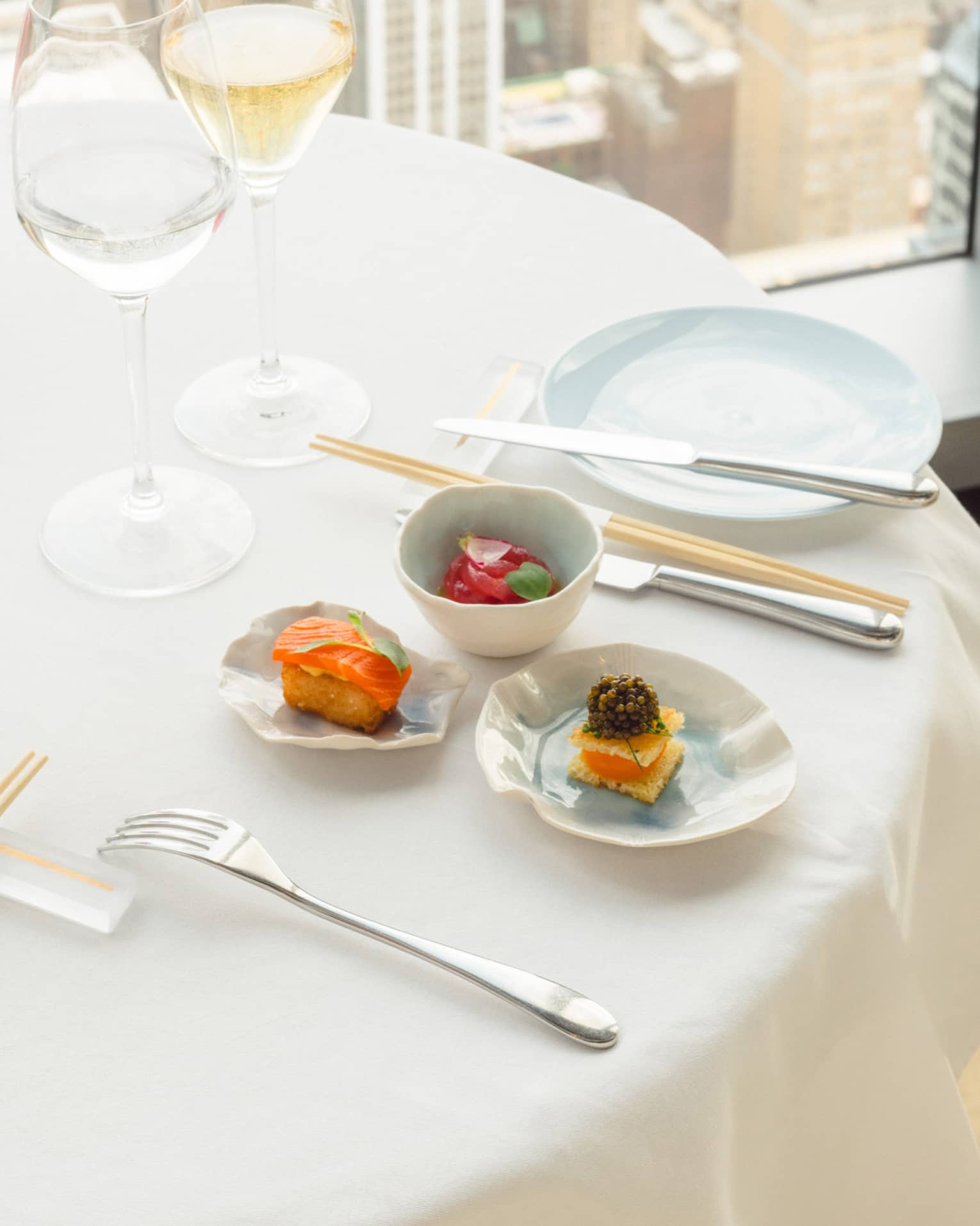 Three small, artfully designed bites of food with wine on an elegantly set table; a city far below is glimpsed out a window.
