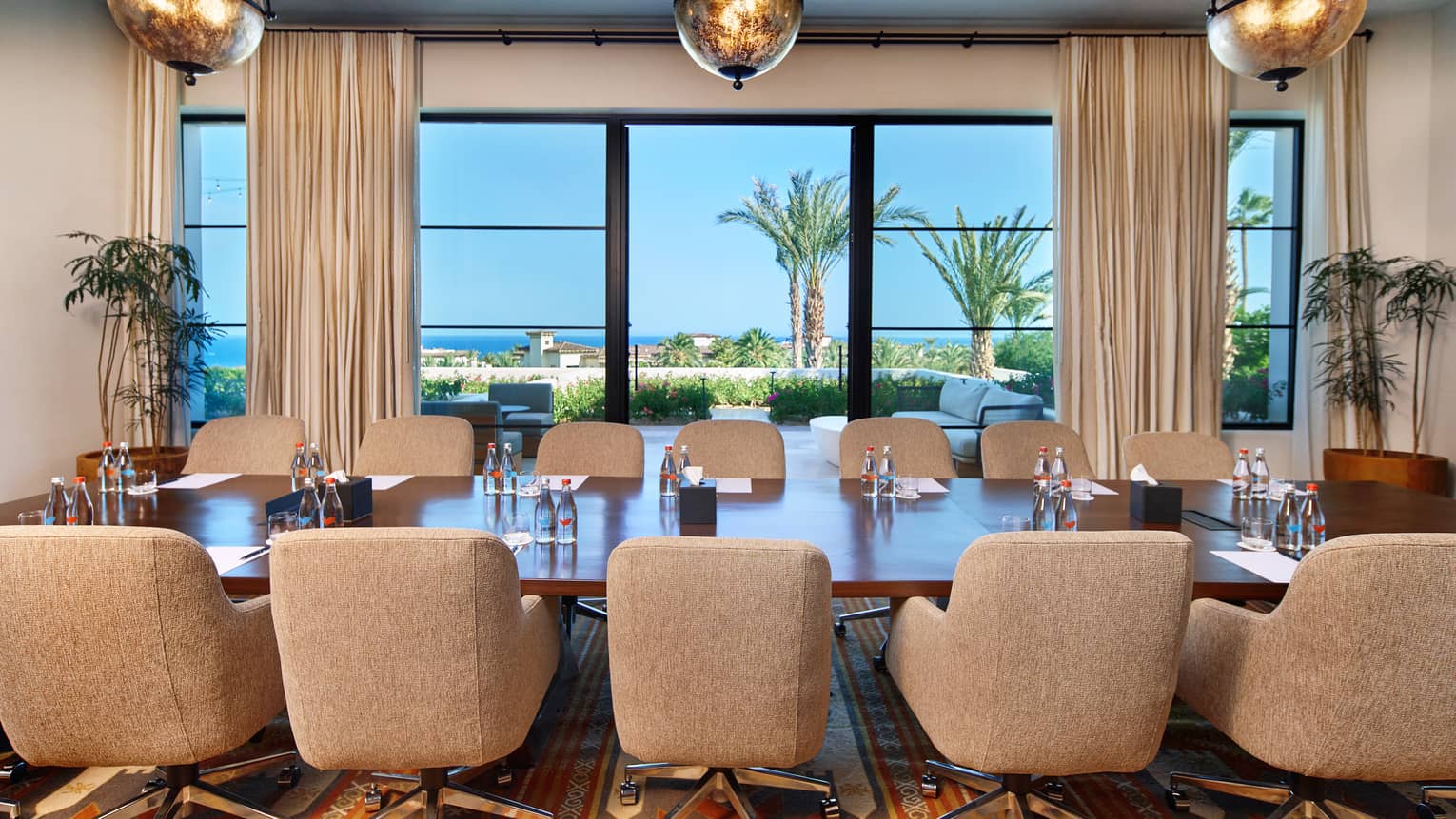 Upholstered seating and a long meeting table in a spacious room overlooking views of palm trees and the ocean. There are globe-shaped pendant lights over the table, room accents of potted greenery, and neutral-colour decor.