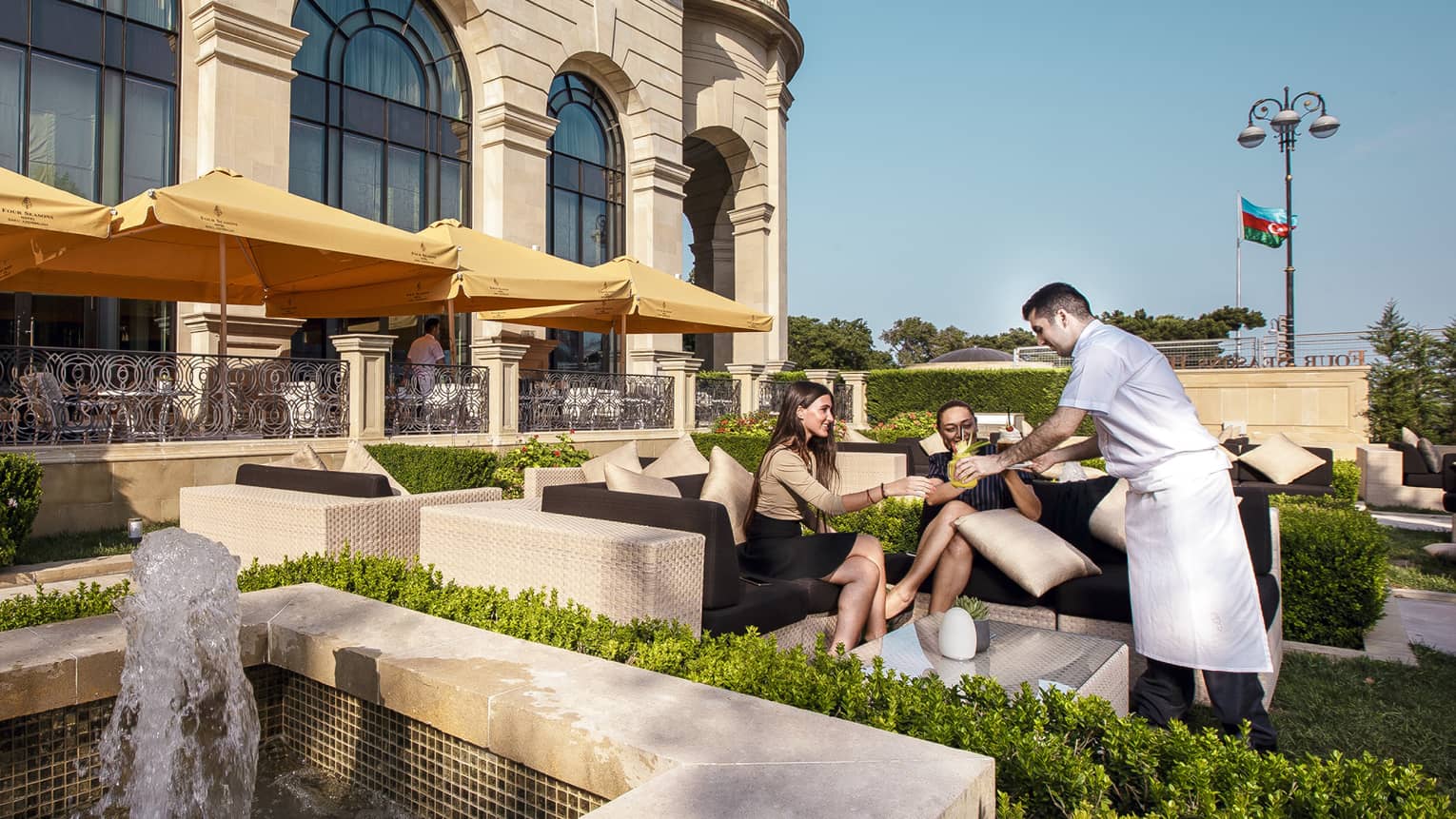 Hotel staff serves cocktails to two women on wicker sofa in large outdoor patio