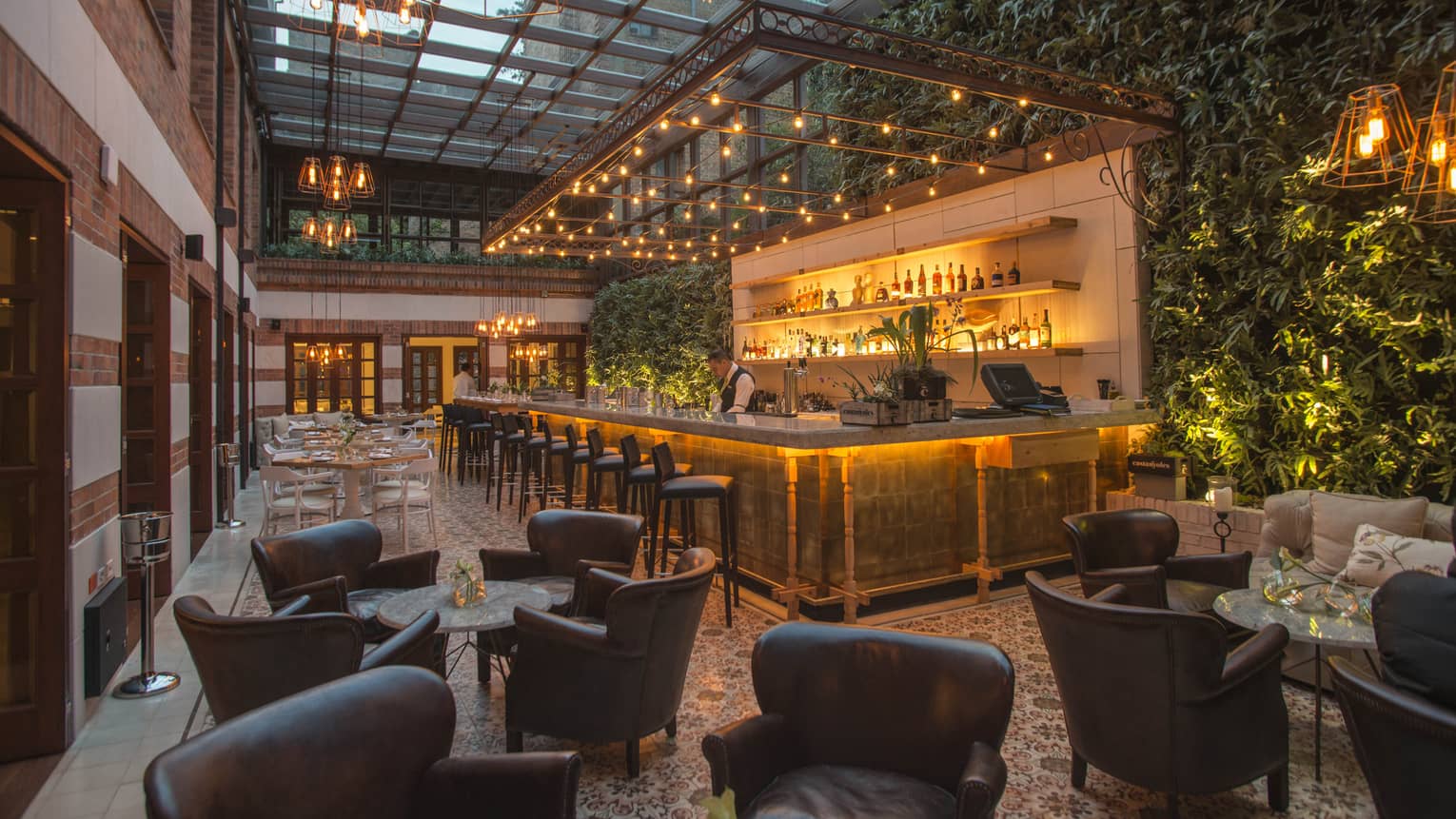 A bar counter surrounded by twinkle lights is set in front of a green wall filled made up of live plants, with numerous round cocktail tables and brown leather club chairs in the foreground 