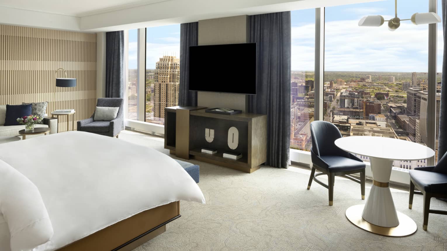 A hotel room with large windows on one wall, a countertop, a television, a small table with two chairs, a bed, and sofa.