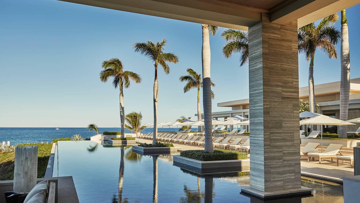 Infinity pool with potted palm trees next to row of white lounge chairs, umbrellas, ocean view