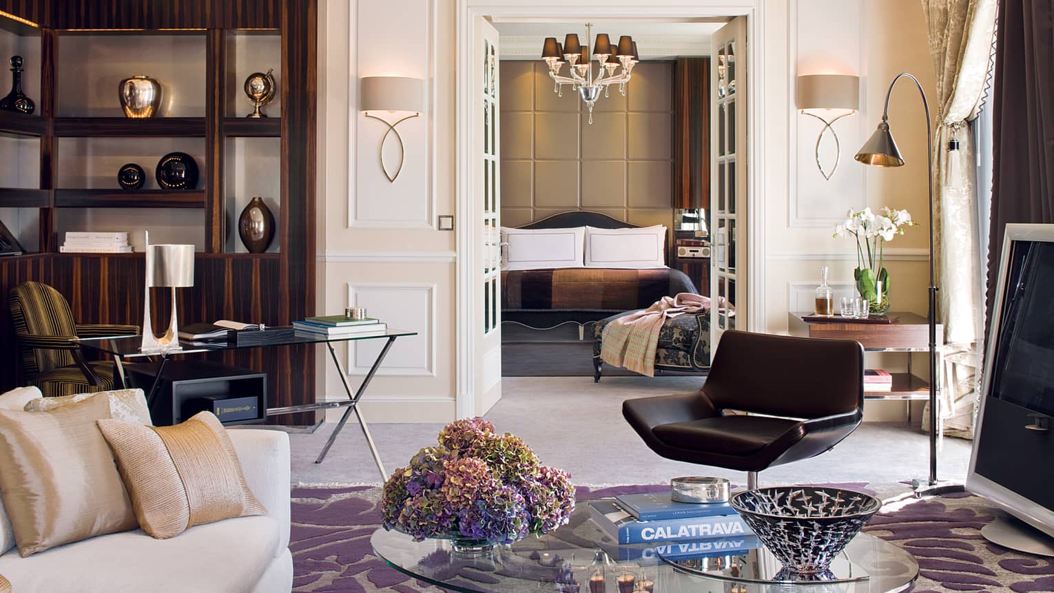 Hotel living room with coffee table, book, retro black chair, glass desk in front of shelf, vases, bedroom door