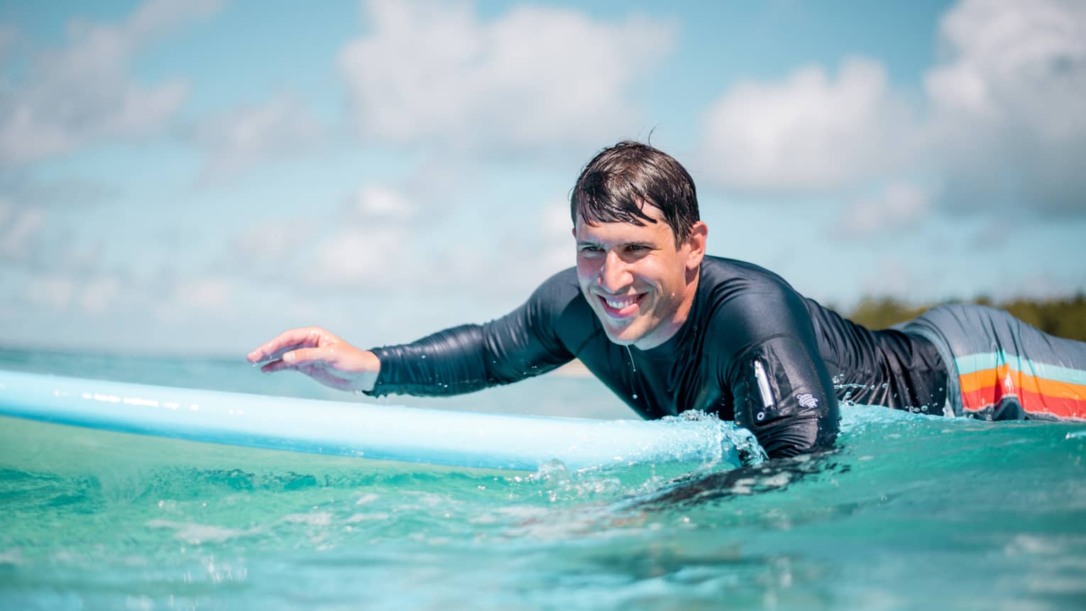 Man in rash guard smiling while paddling on blue surfboard in blue-green water